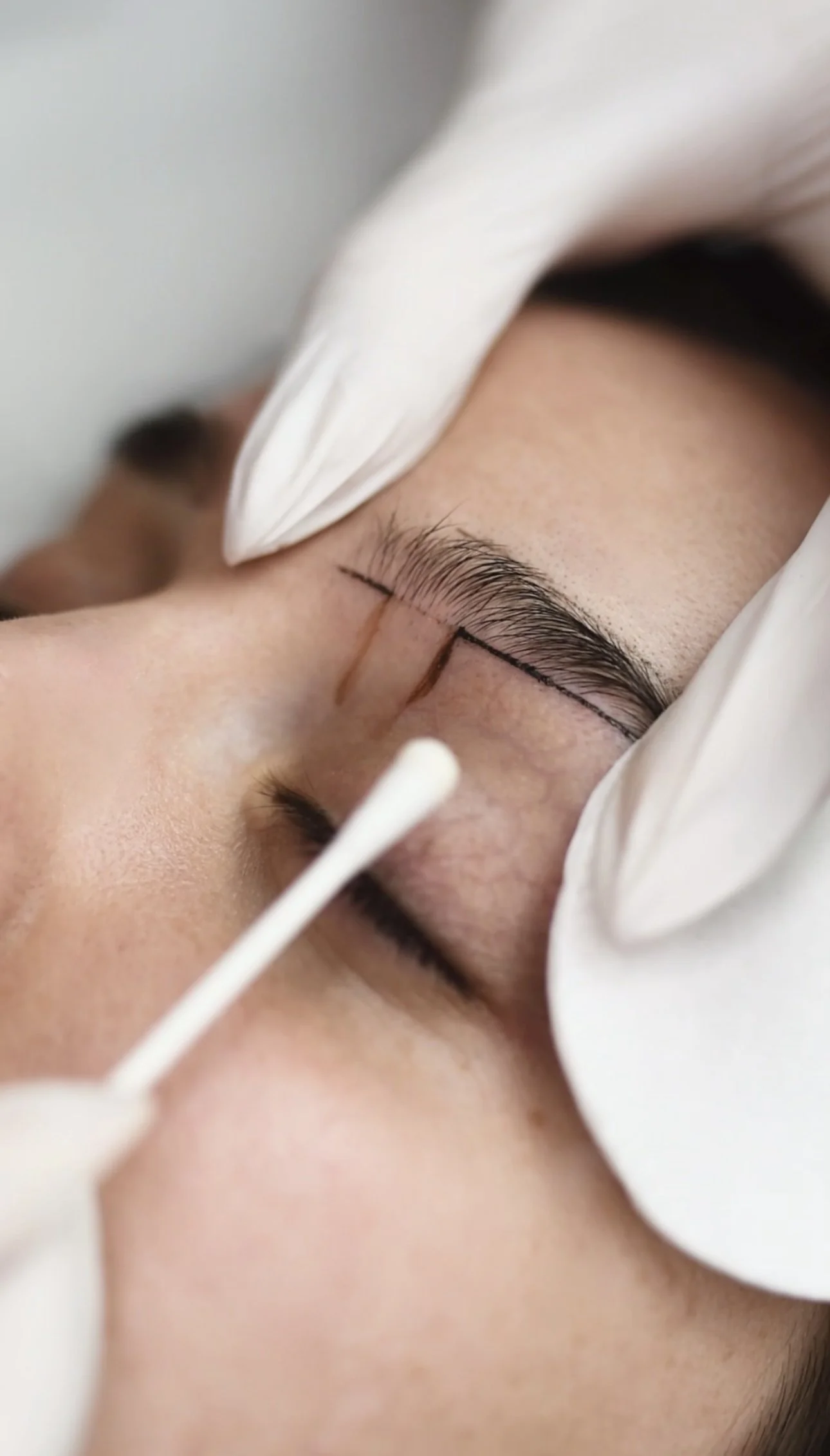 Close-up of a person receiving a cosmetic eyelash treatment, with markings on the eyelid and a cotton swab near the eye.