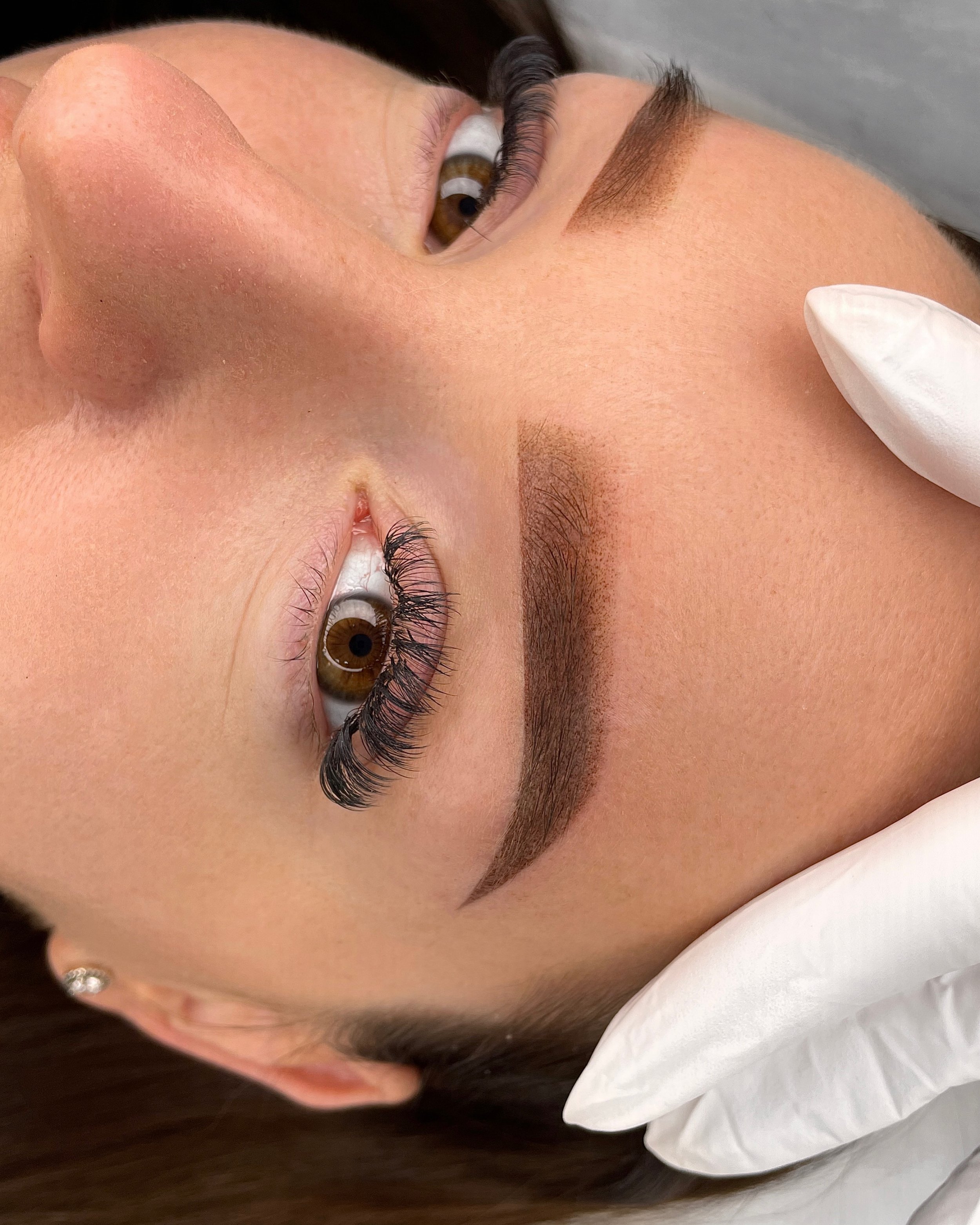 Close-up of a woman’s face showing freshly done eyebrow and eyelash extensions, with a gloved hand gently supporting her head.