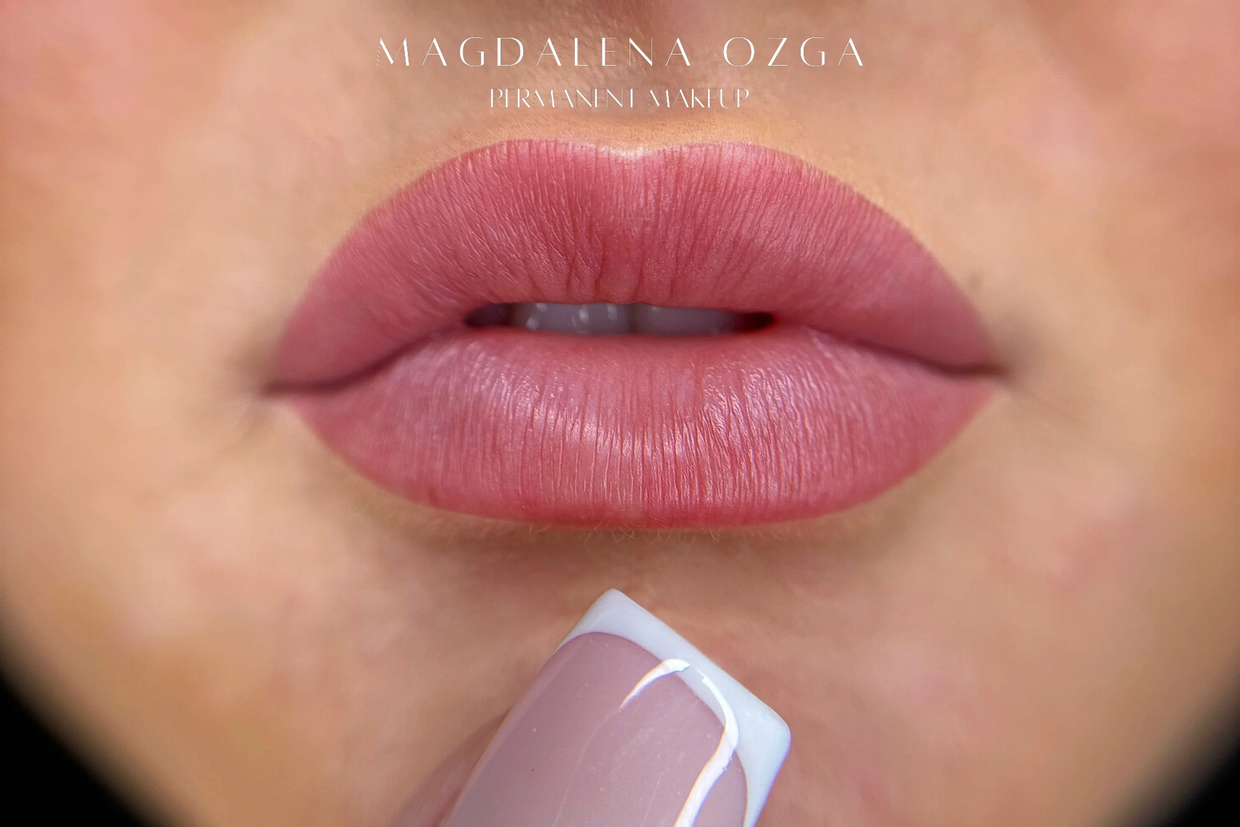Close-up of a person's lips with pink matte lipstick, with a hand applying lipstick or lip balm. Text overlay reads 'Magdalena Ozga Permanent Makeup'.