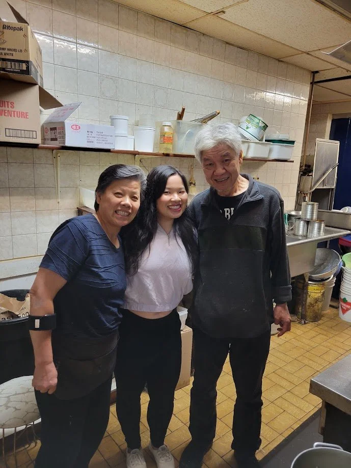 Two smiling women and a smiling man pose for a photo inside a large kitchen.