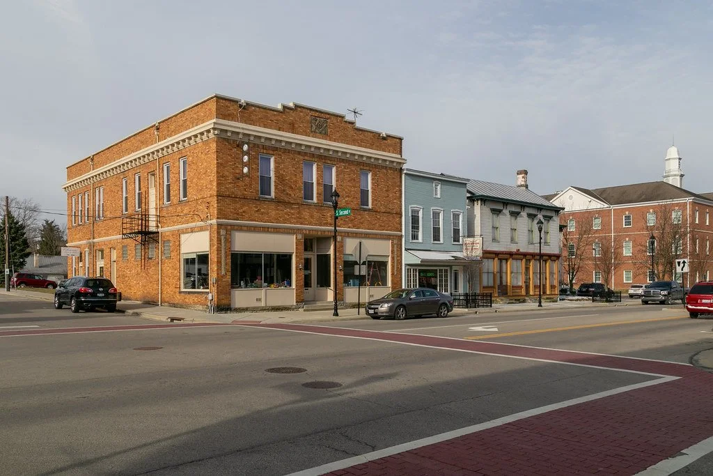 A street view with a corner brick building and several smaller storefronts, parked cars, and a traffic sign under a cloudy sky.