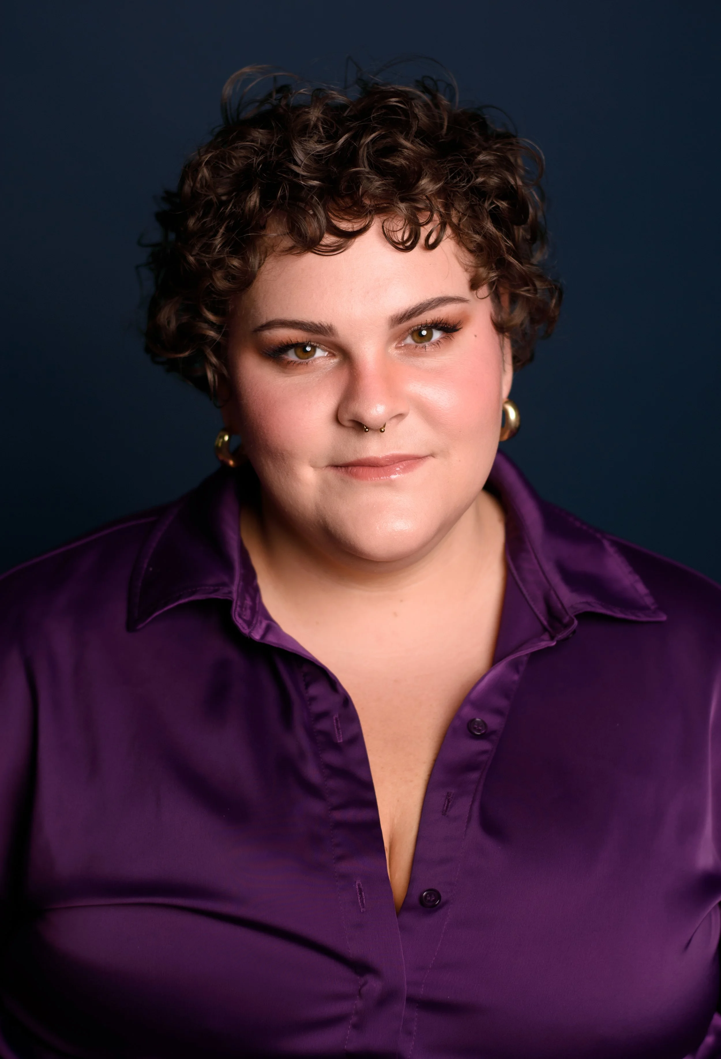 Portrait of a woman with short curly brown hair, wearing a purple satin shirt and gold jewelry, against a dark background.