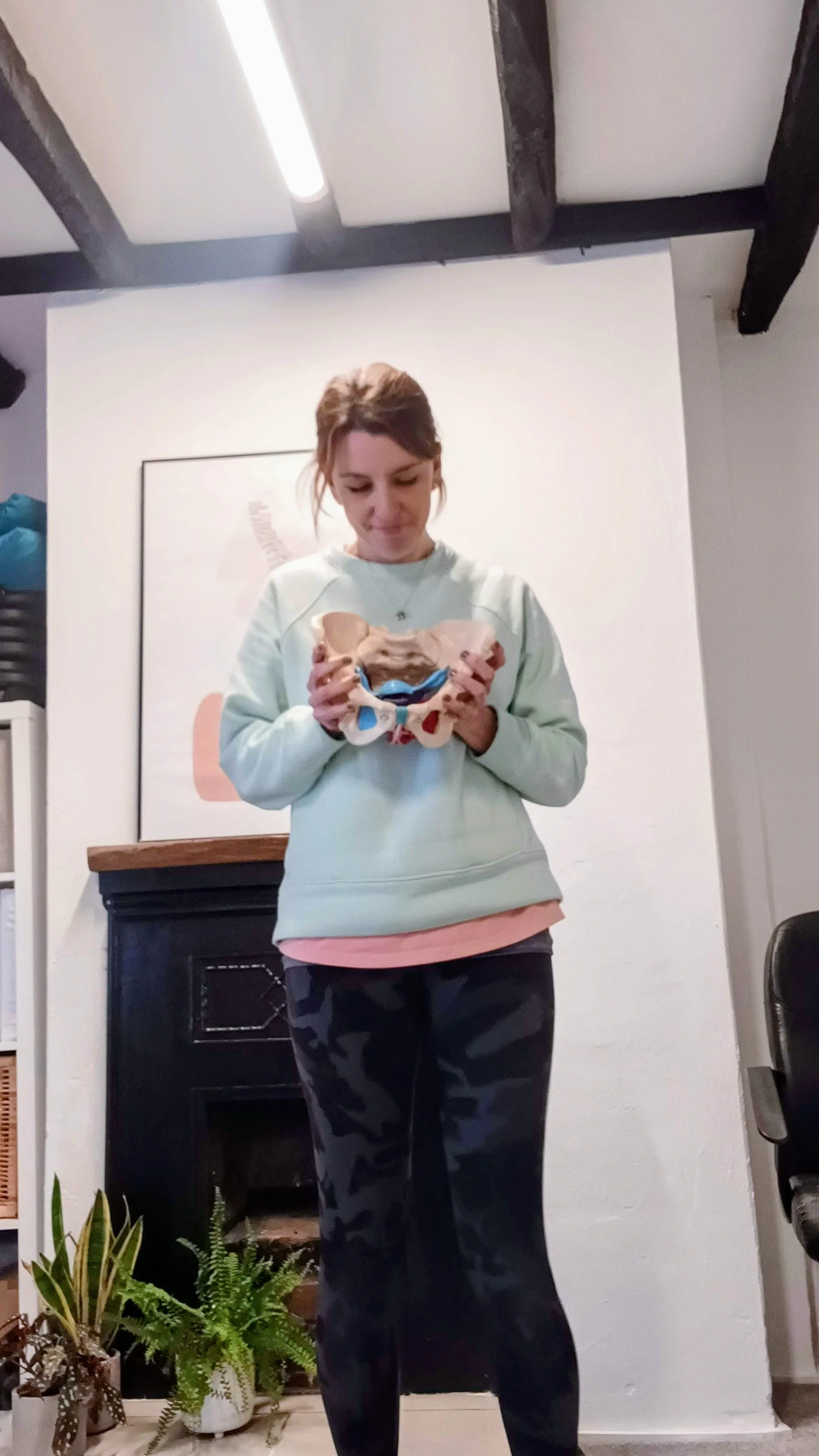A woman standing indoors, holding a model of a human pelvis and displaying it while looking down. She is wearing a light sweatshirt with a pink shirt underneath and dark camouflage pants. There are plants at her feet, a black fireplace behind her, and a framed picture on the wall.