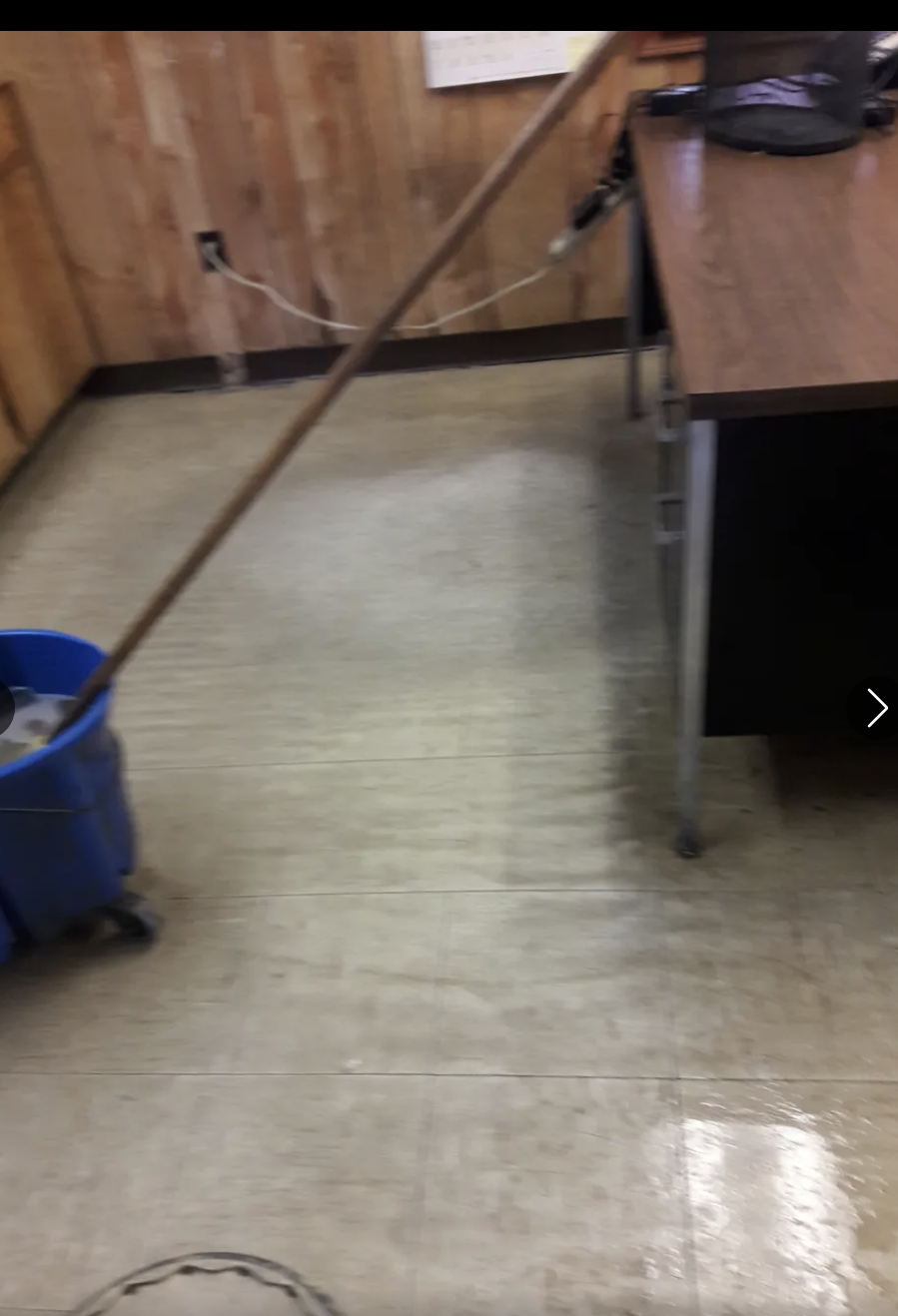 Office space with wooden paneled wall, a desk with a black chair, a blue bucket, and a mop handle leaning against the desk.