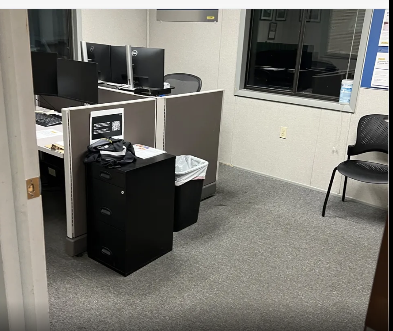 An empty office room with several computer workstations, a black filing cabinet with clothes on top, a trash can, and a single black chair near the window.