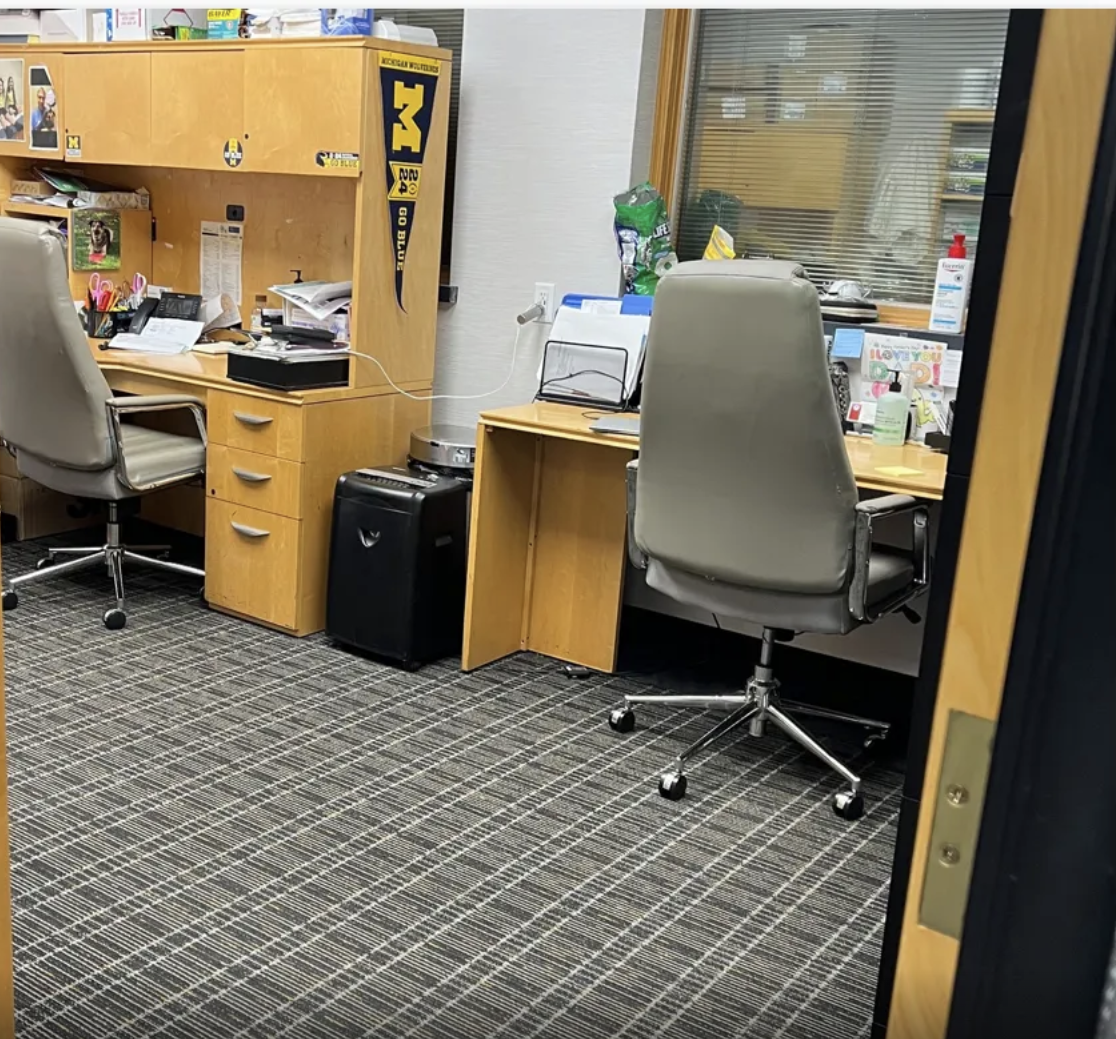 Office with two desks, office chairs, computer, paperwork, and office supplies, viewed through an open door.