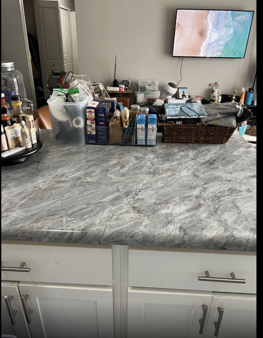 Kitchen countertop with various food and household items, including jars, boxes, and a plastic container, with a cluttered background featuring a TV, picture frame, and miscellaneous objects.