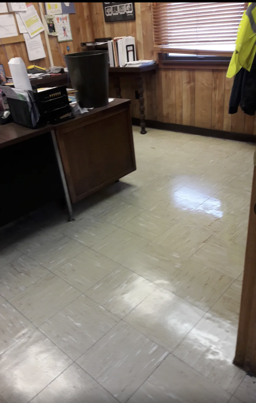 Office or workspace with wood-paneled walls, a desk cluttered with papers and office supplies, a large black trash can, and a window with wooden blinds, with a coat hanging on the wall.