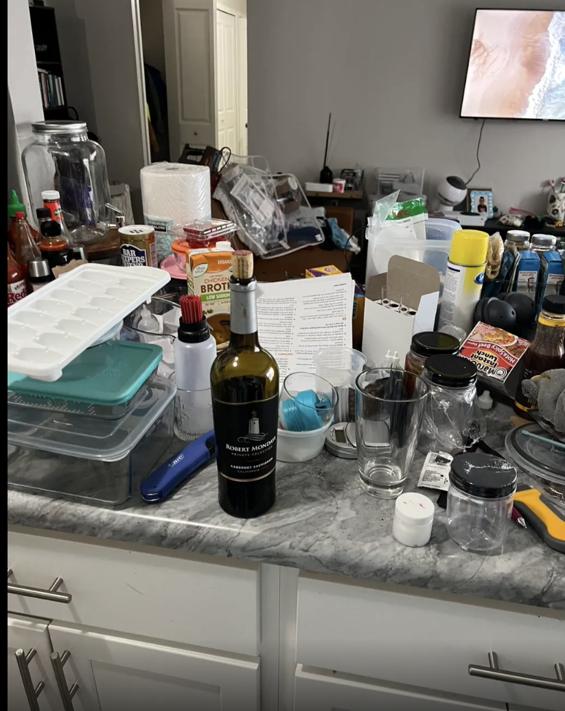Messy kitchen countertop with wine bottle, glasses, food containers, paper towels, and various kitchen items.