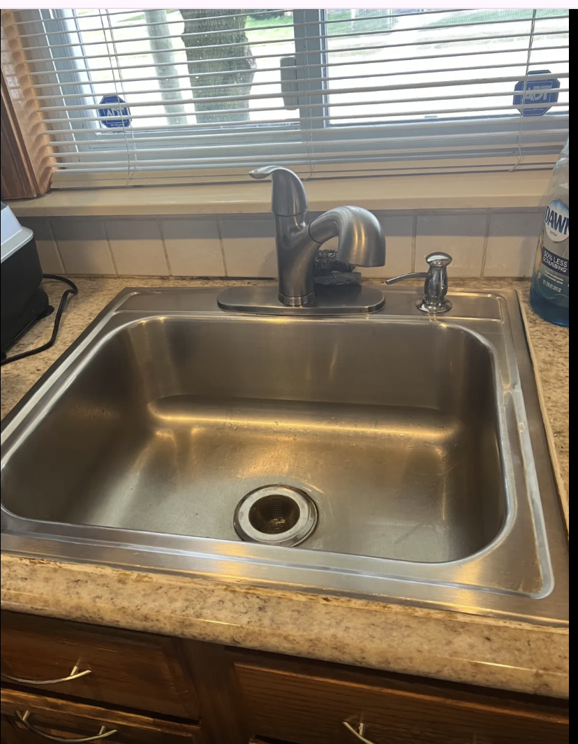 Stainless steel kitchen sink and faucet on a granite countertop below a window with blinds, with a bottle of Dawn dish soap on the right.
