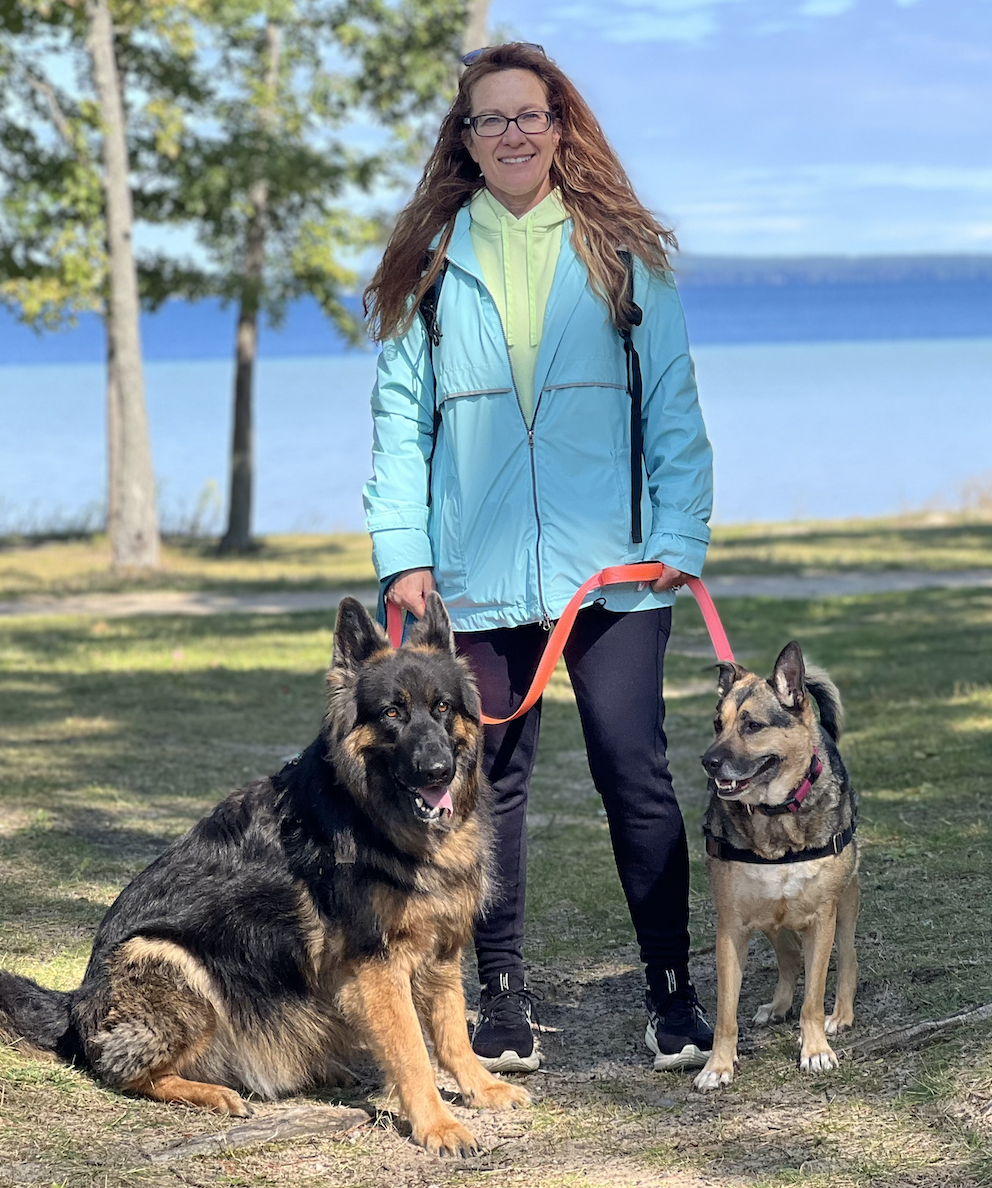 A woman with long red hair, glasses, and a bright blue windbreaker stands outdoors on a grassy area near a body of water, smiling at the camera. She holds leashes attached to two dogs, one sitting in front of her on the left and the other standing to her right. The dog on the left is a large German Shepherd with black and tan fur, and the dog on the right is a smaller mixed breed with tan and black fur. In the background, there are trees, a lake, and a partly cloudy sky.