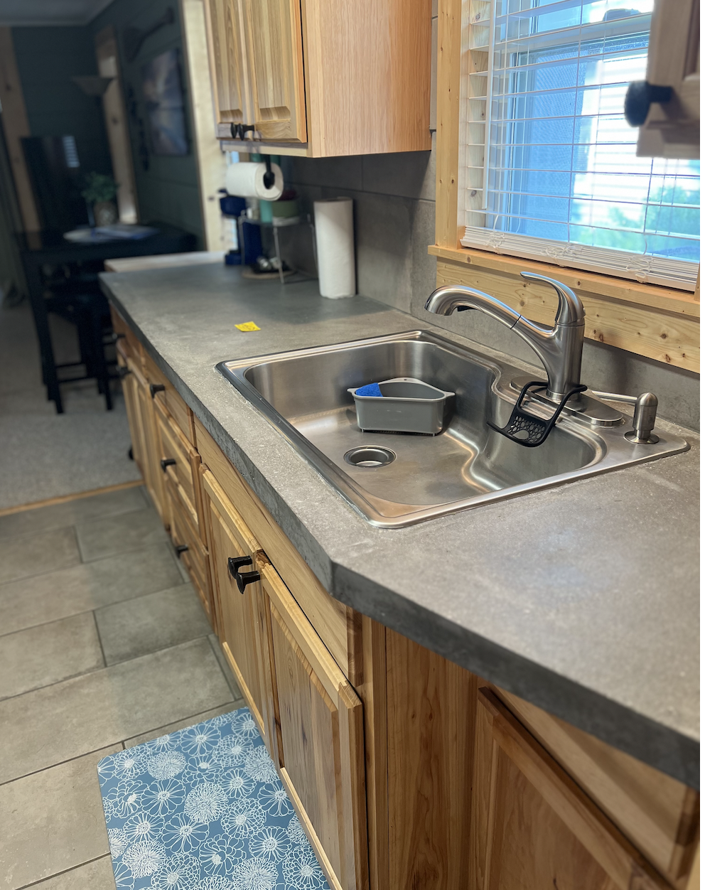 Kitchen countertop with a stainless steel sink, faucet, sponge, and dish soap holder, wooden cabinets, window with horizontal blinds, and a blue floral-patterned rug on tile floor.