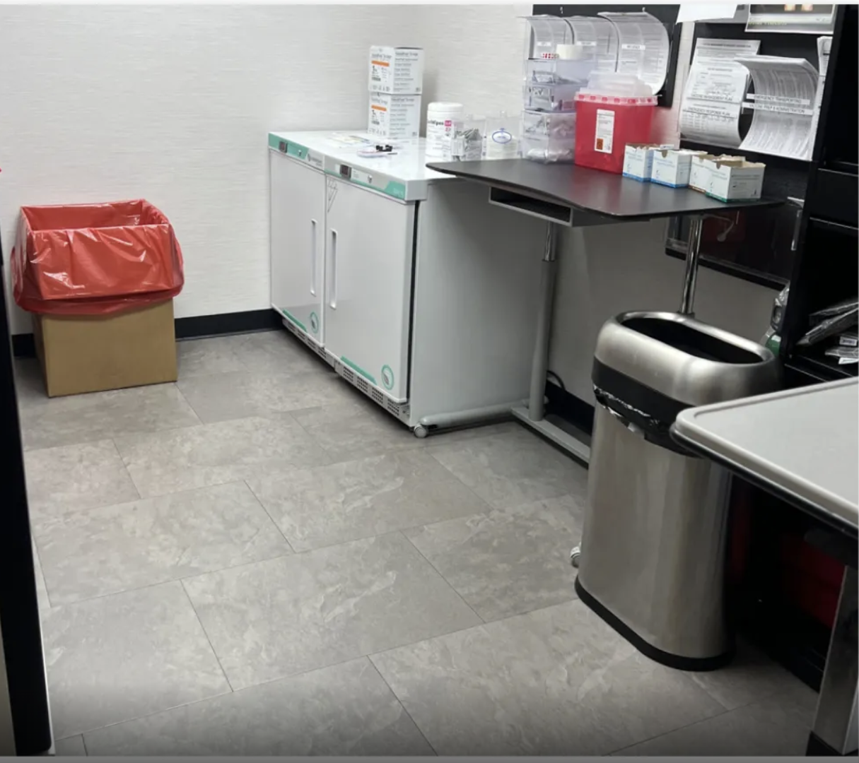 Empty medical supply room with a red trash bin, a gray cabinet, a black counter with boxes and bottles, and a stainless steel trash can.