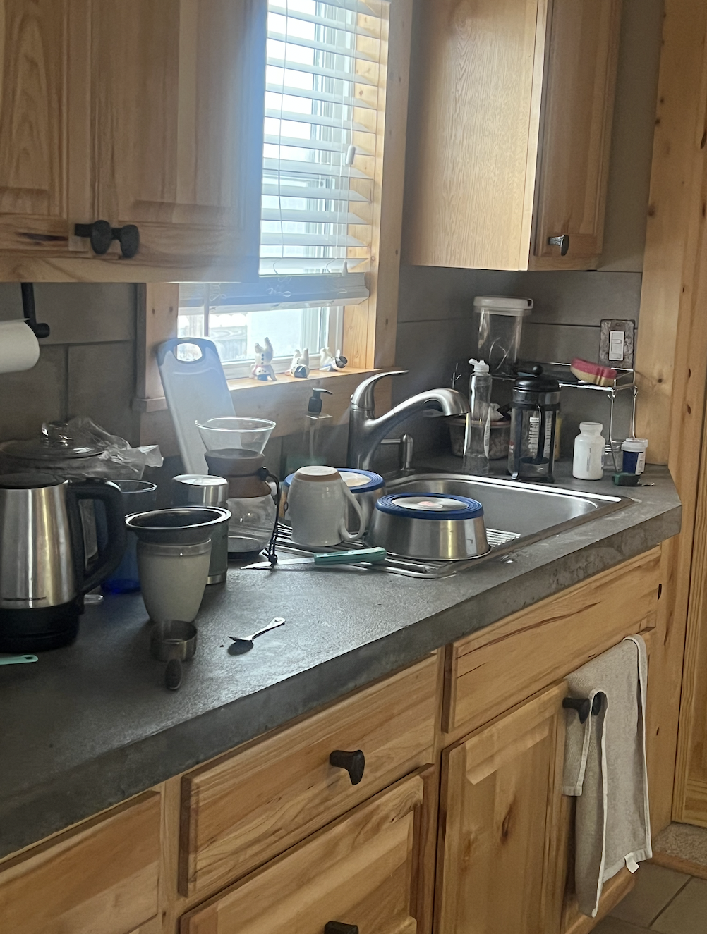 Cluttered kitchen sink area with dishes, cups, and utensils, adjacent to a window with blinds and wooden cabinets.