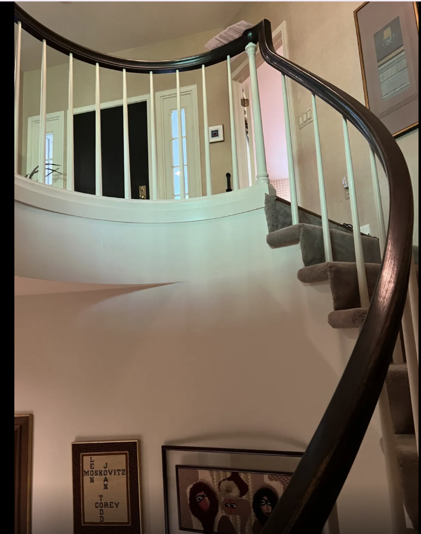 View of a staircase with gray carpeted steps and white railings, leading to an upper floor in a home interior, with framed artwork and a picture frame on the lower wall.