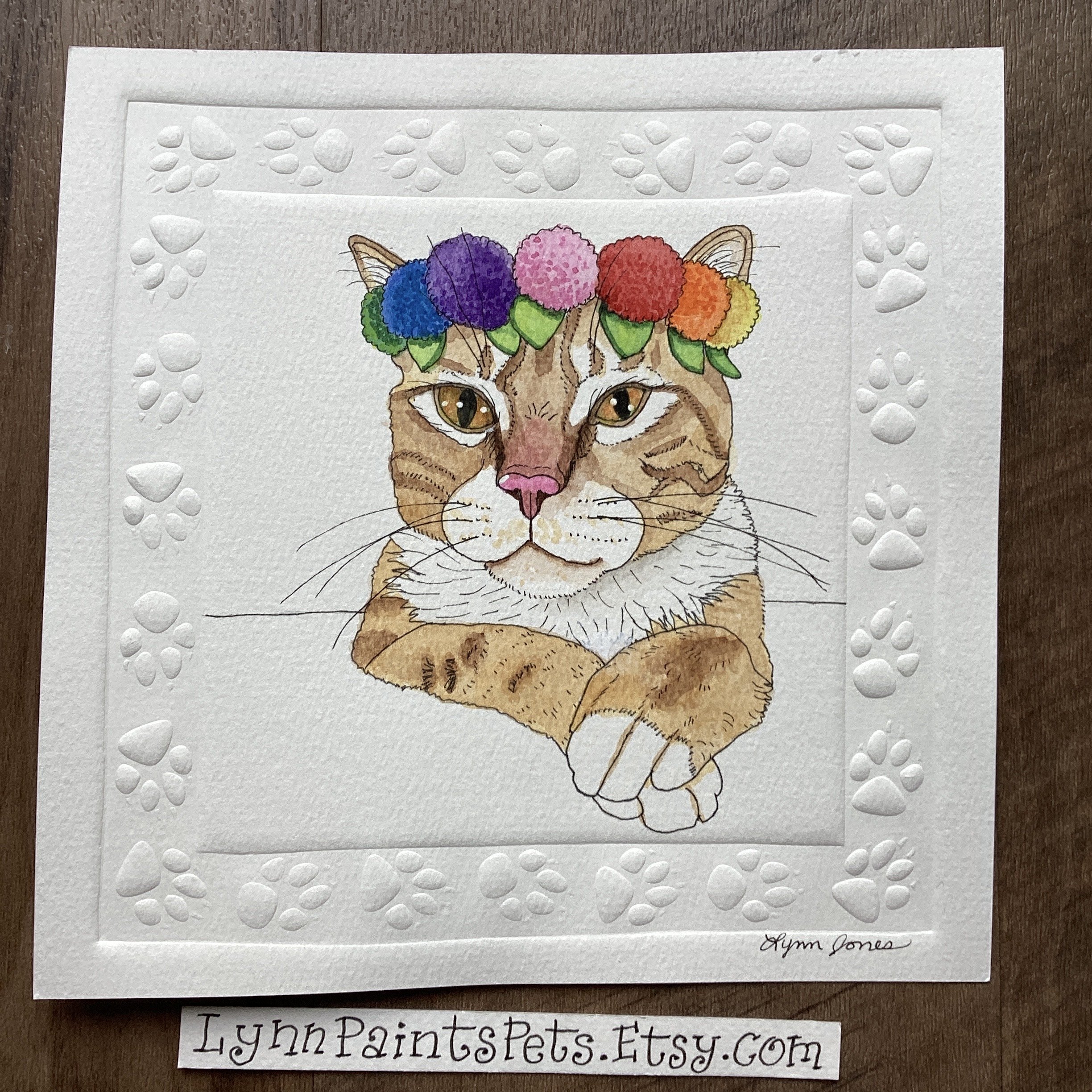Watercolor illustration of a cat wearing a colorful pom-pom floral crown, with its paw crossed in front of it, surrounded by a textured paw print border, signed by artist Hyun Jones.
