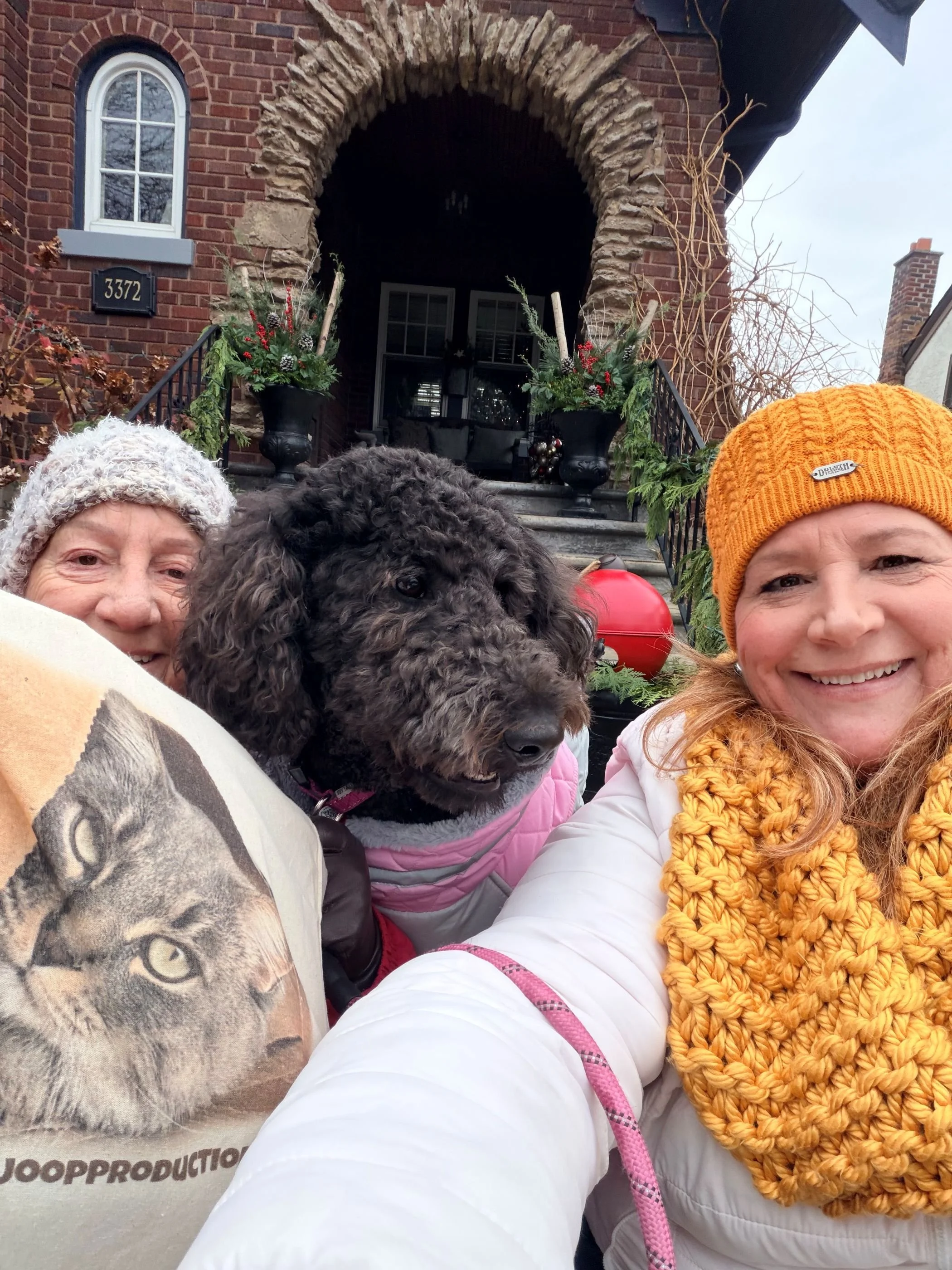 Two women and a dog taking a selfie in front of a brick house decorated for the holidays. The woman on the right is wearing an orange beanie and a yellow scarf. The woman on the left is wearing a gray knit hat. The dog, a curly-coated breed, is wearing a pink jacket and is between them.