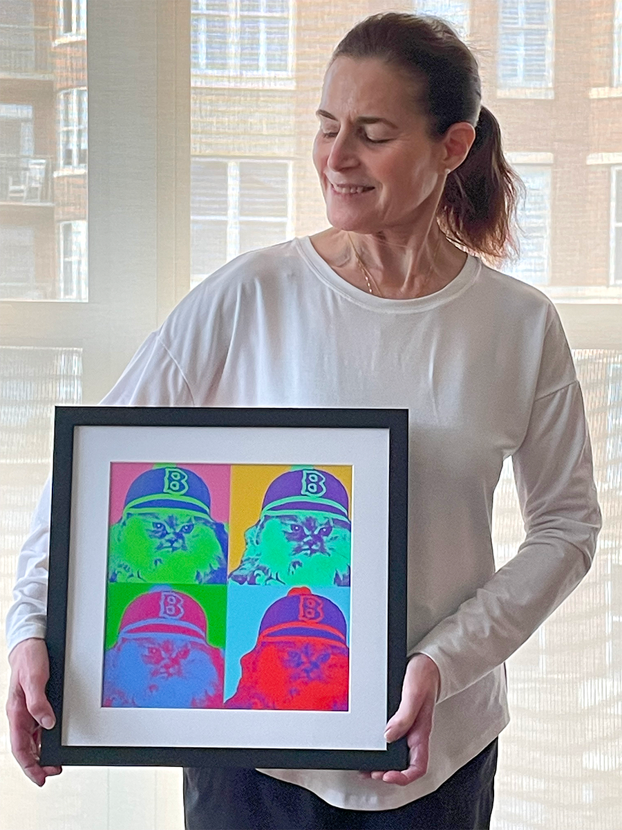 A woman smiling and holding a framed pop art print of a cat wearing a baseball cap in four colorful panels.