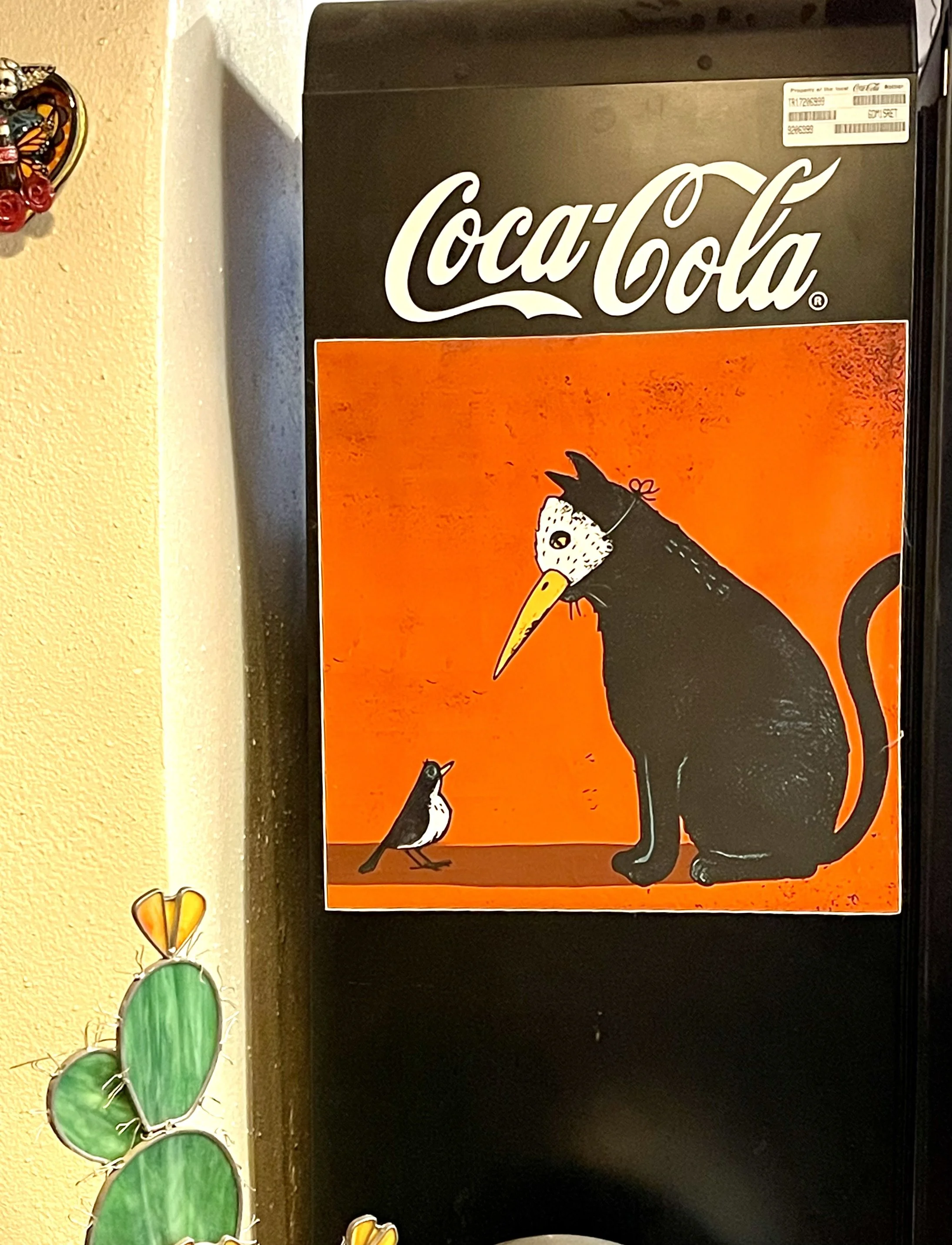 A refrigerator with a Coca-Cola sticker featuring an illustration of a cat with a beak and a bird facing each other against an orange background.