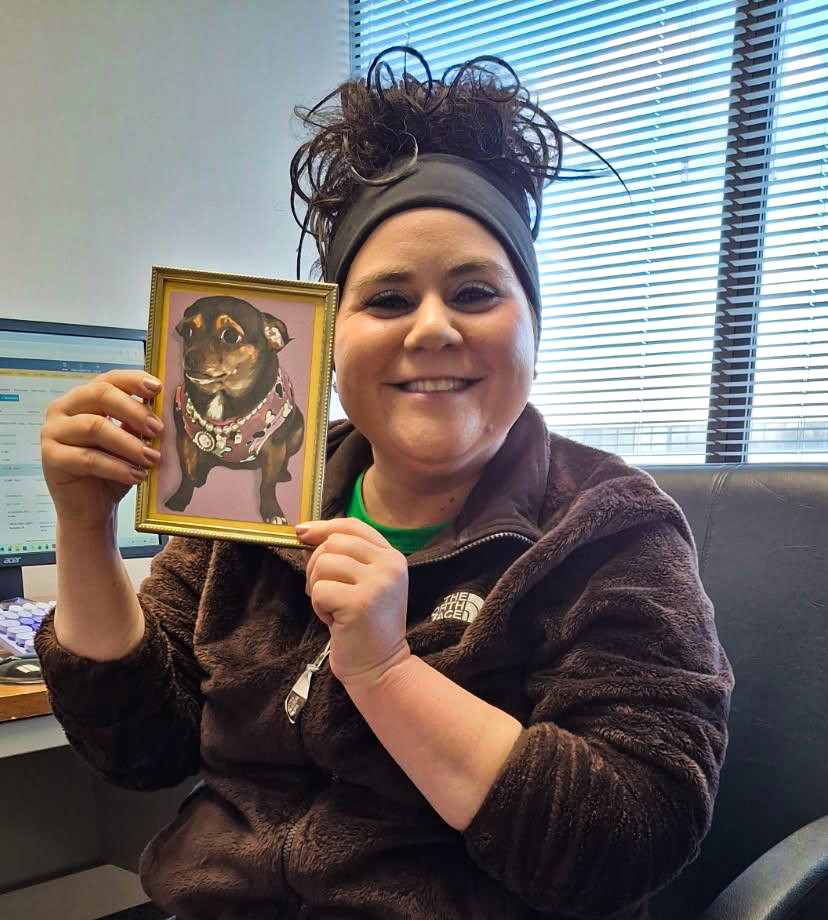 A woman with dark curly hair in a ponytail, wearing a black headband and a brown fleece jacket, smiling and holding a framed picture of a small dog with a pink background, at an office desk with a computer and window blinds in the background.