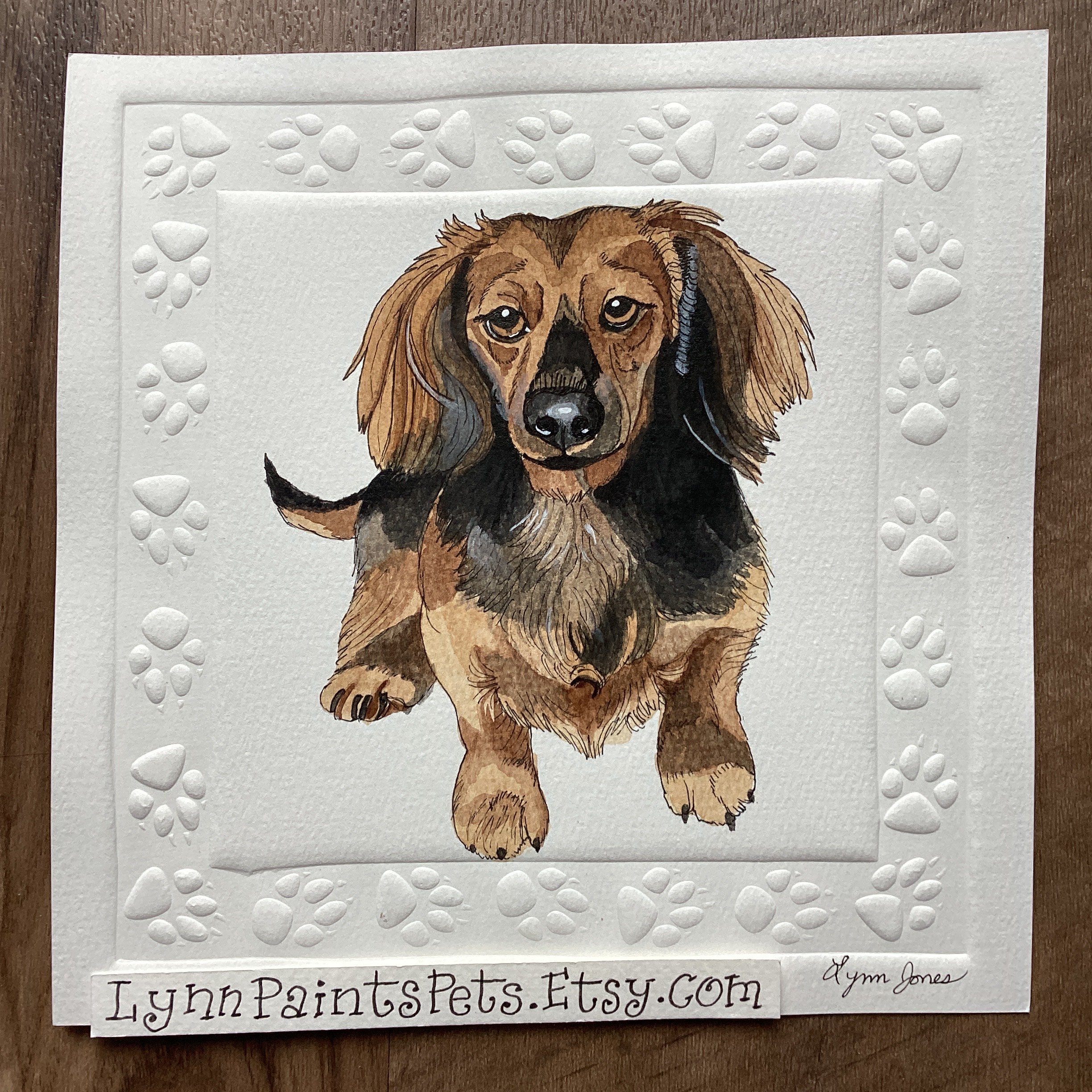 A watercolor painting of a brown and black dachshund puppy on white paper with embossed paw print border. The artwork is signed by Lynn Jones and has the website LuzzyPaintsPets.Etsy.com written at the bottom.