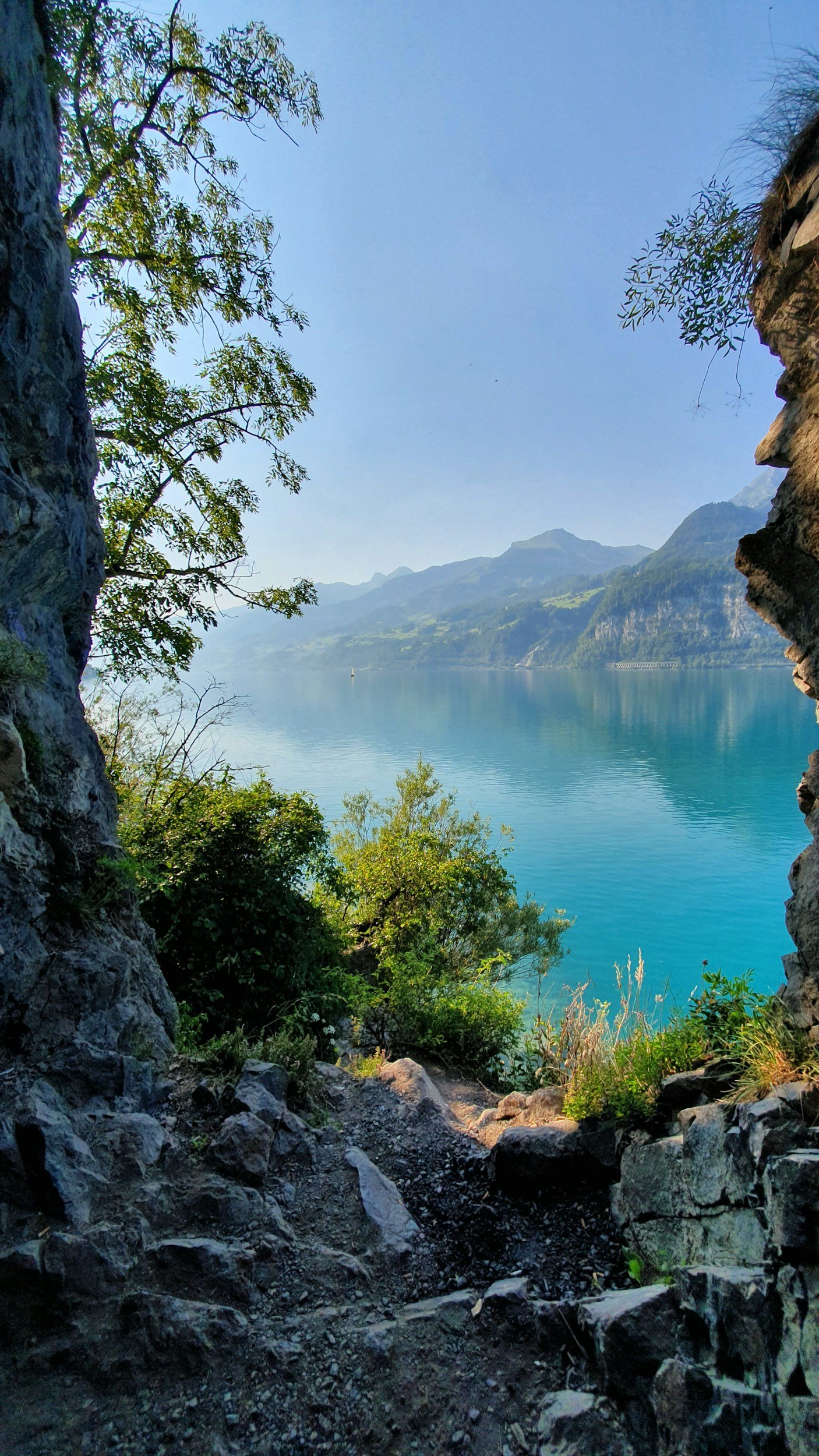 View through rocky cave opening overlooking a blue lake with green hills and mountains in the distance, under a clear sky.