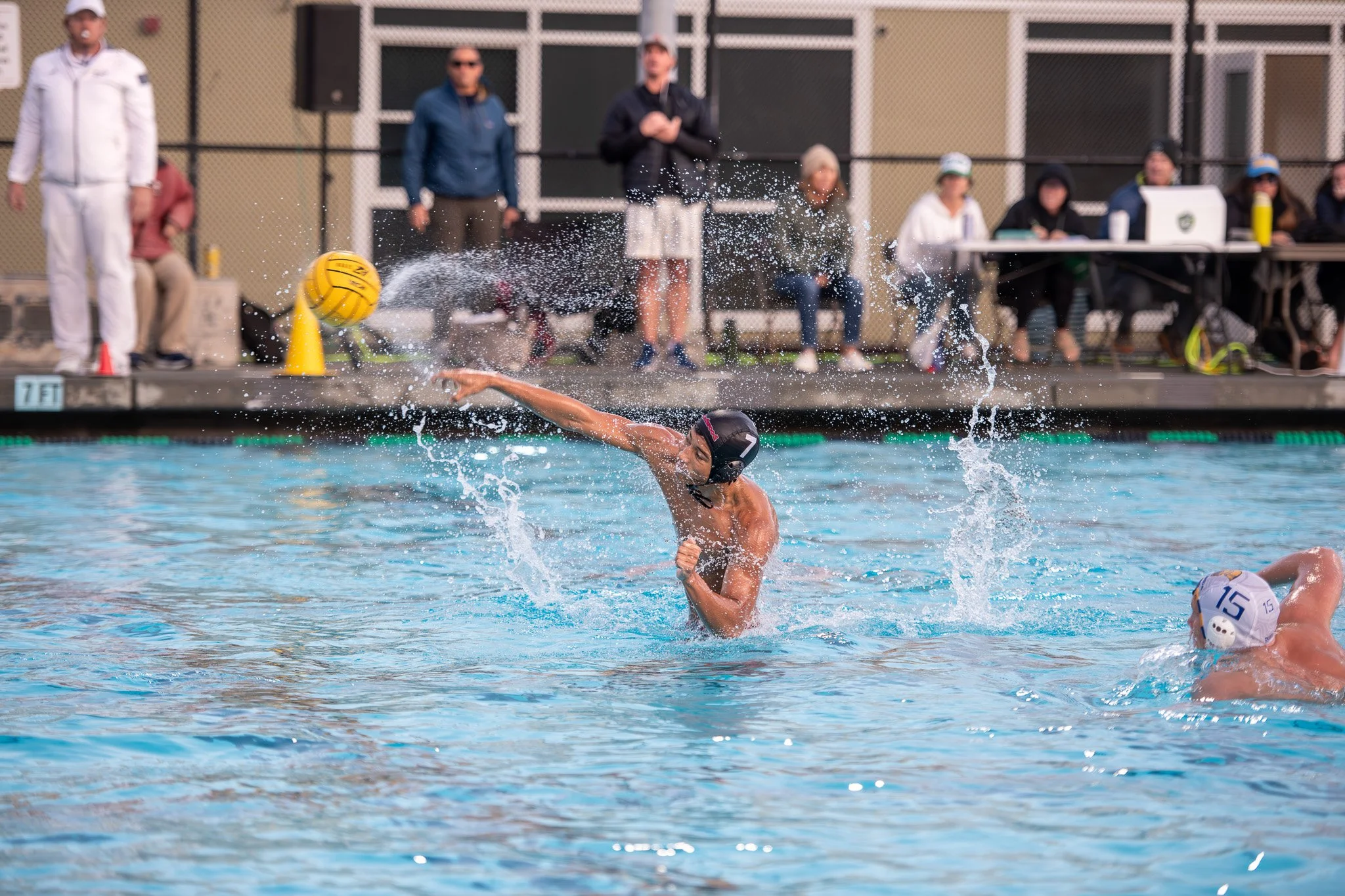 A water polo game with players in the pool, one player in a black cap is throwing the yellow ball, another player wearing a white cap is swimming nearby, and spectators and officials are on the pool deck in the background.