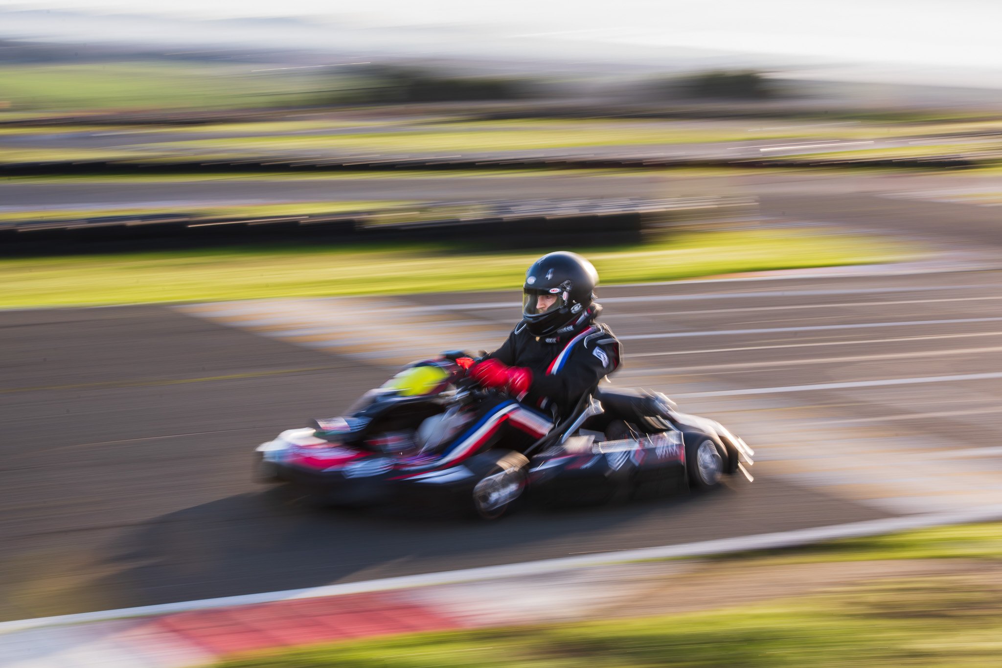 Person in black racing suit and helmet riding go-kart on race track, motion blur background.