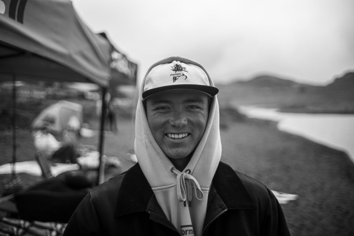 A young man smiling outdoors, wearing a hoodie and a cap, with a blurred tent and landscape in the background.