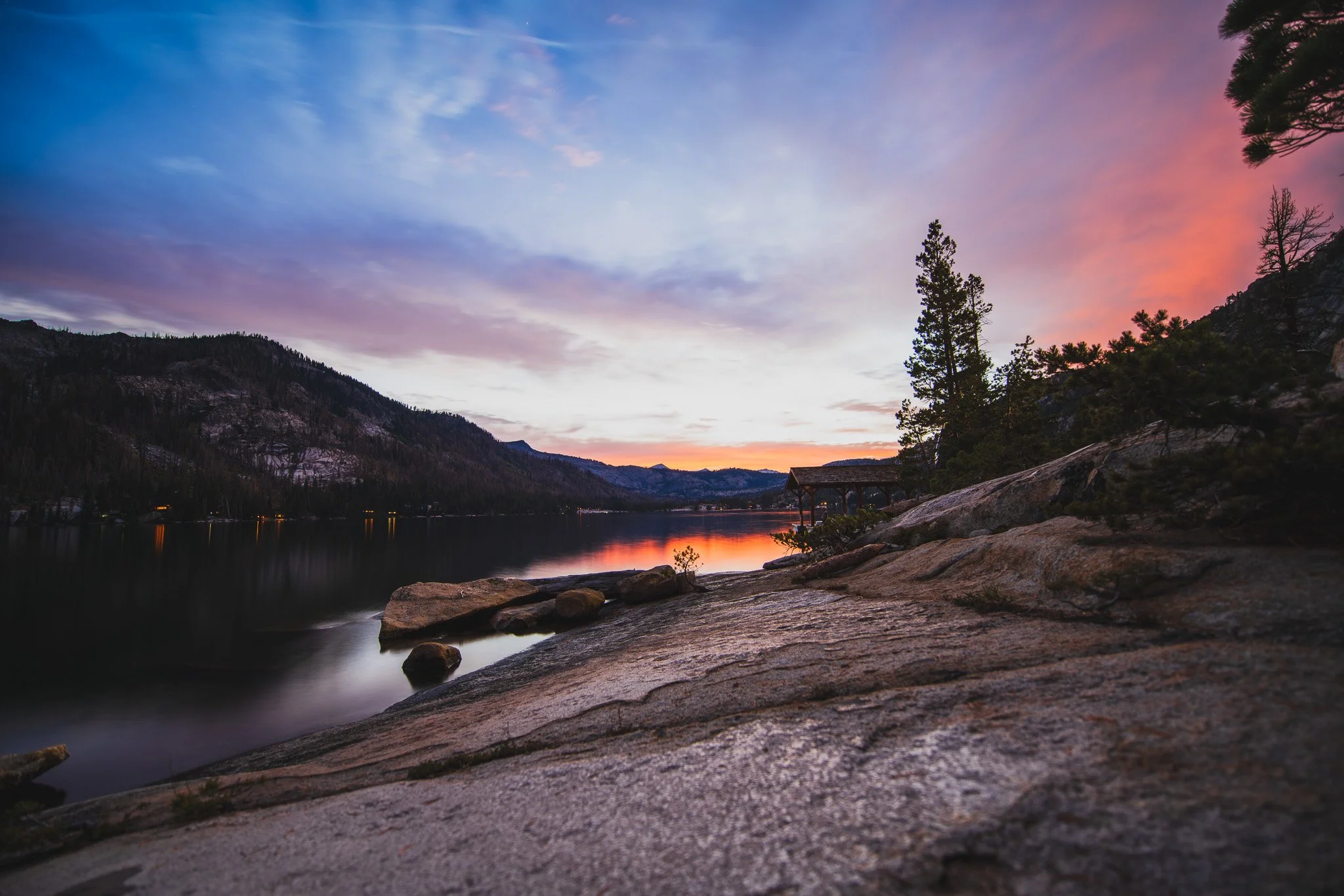 Scenic lakeside view at sunset with colorful pink, purple, and blue sky, mountains in background, and rocky shoreline with trees and a wooden pavilion.