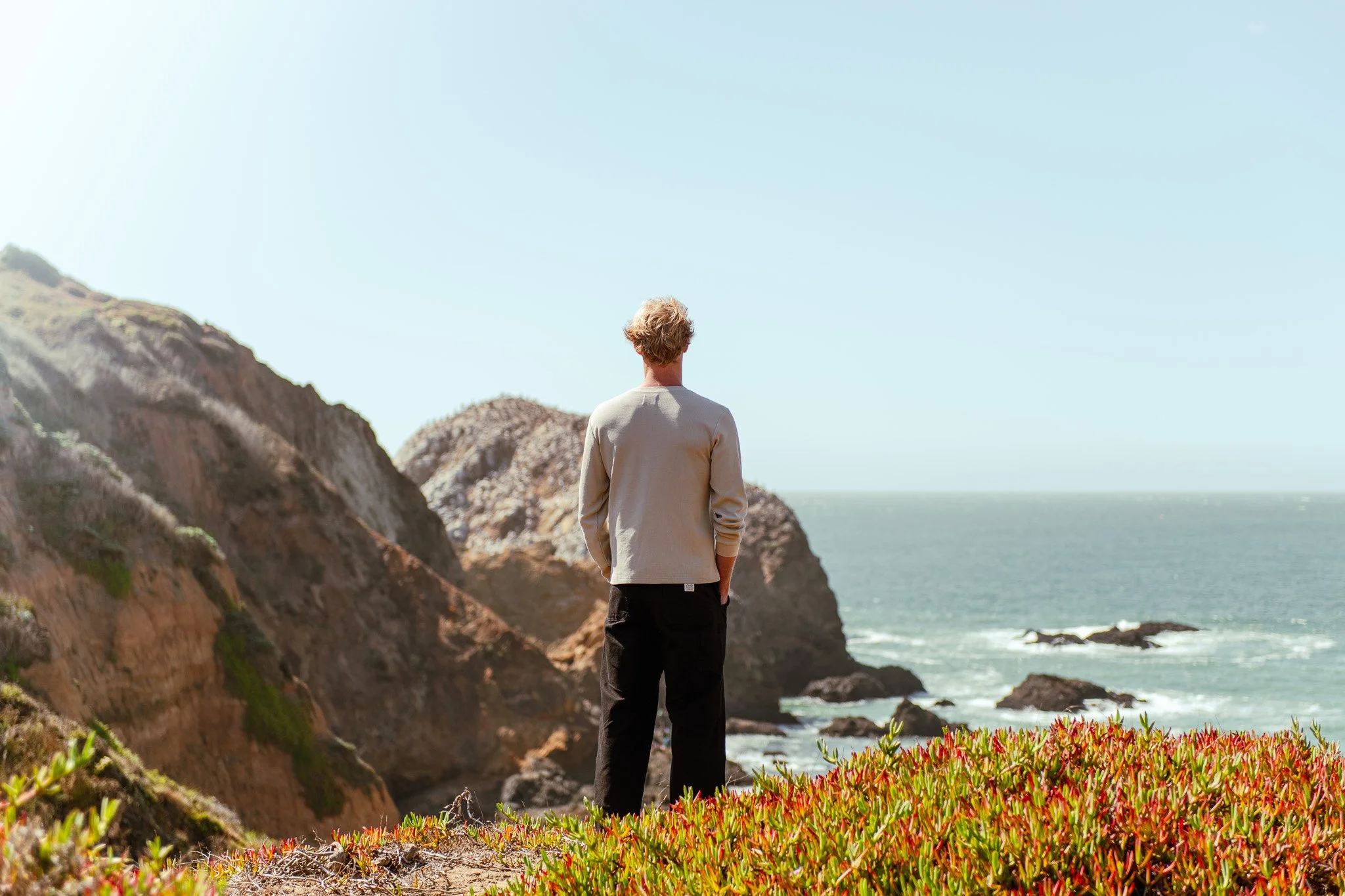 A person standing on a coastal cliff, looking towards the ocean with rocks and cliffs in the background on a sunny day.