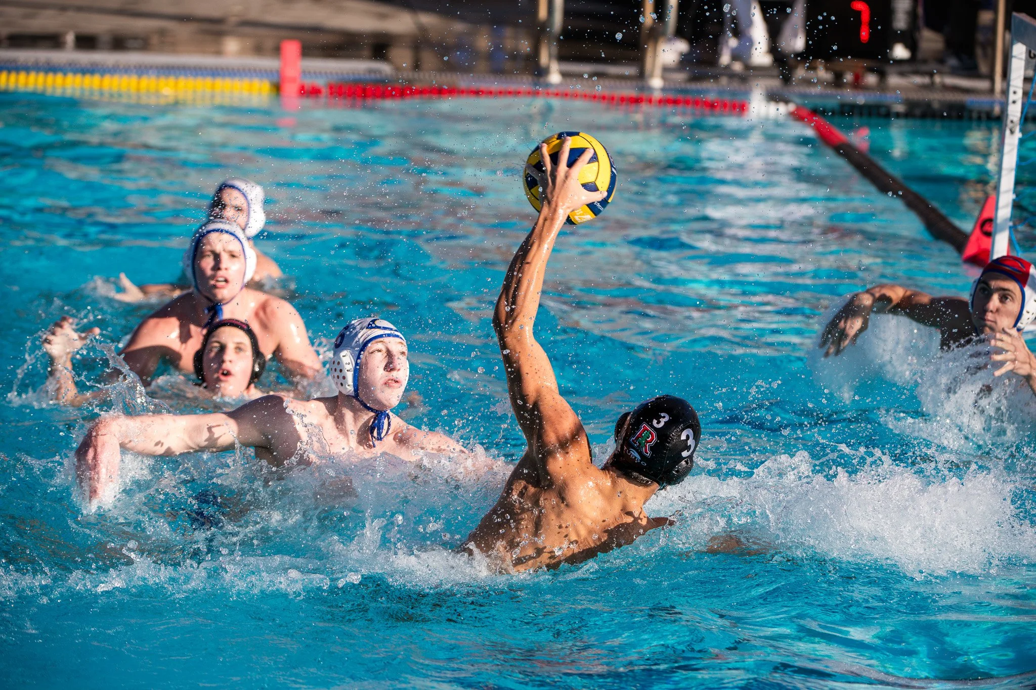 Water polo players competing in a game, with one player reaching out of the water holding a yellow and blue ball, while others swim and attempt to block, wearing caps with protective ear flaps.