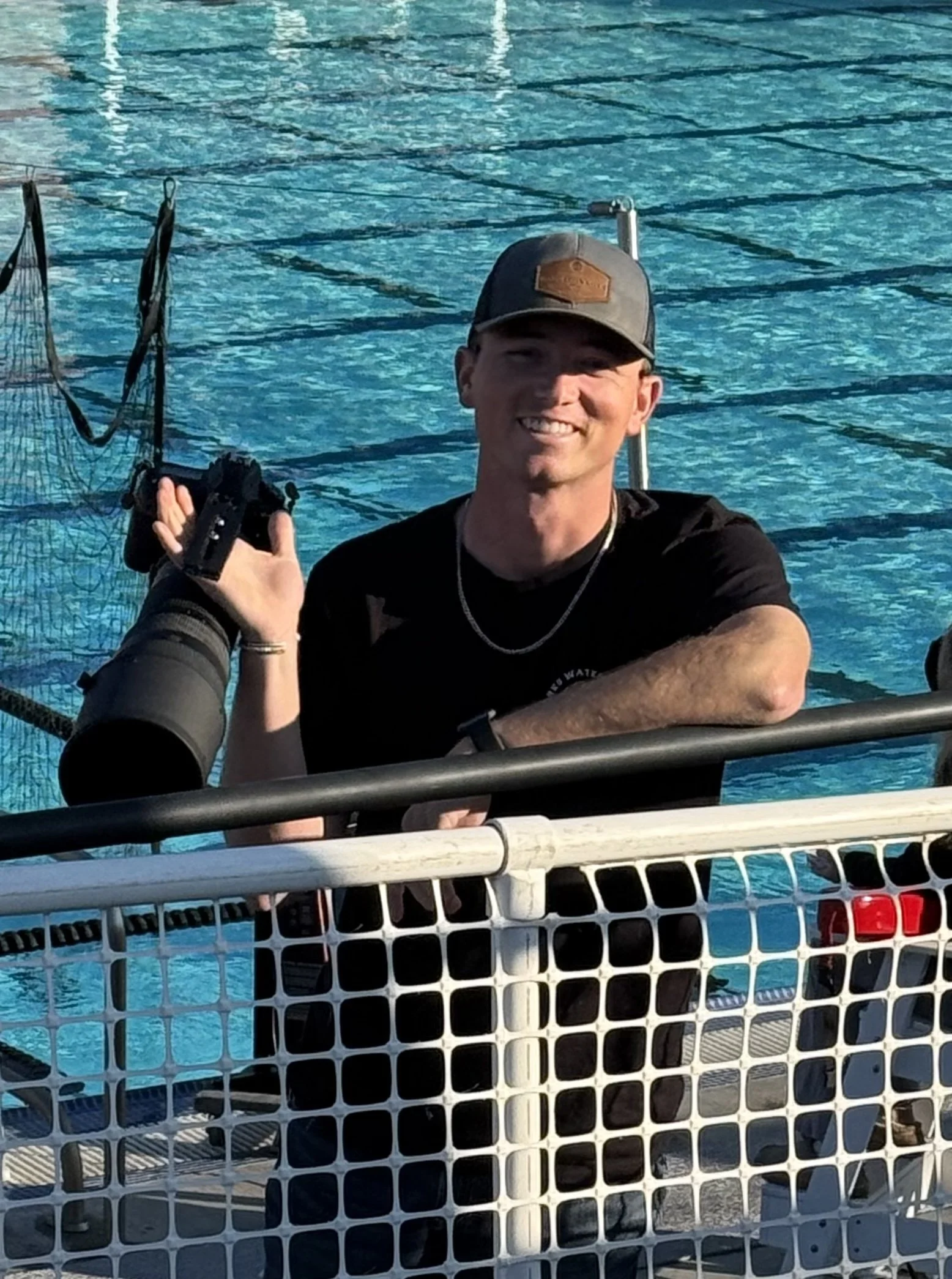 Smiling man with a camera in hand and a cap, leaning on a boat railing near a swimming pool.