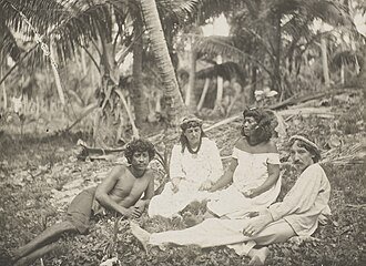 Stevenson and his wife lounge on the grass next to Nan Tok and Natakanti. Stevenson wears  loose pants and shirt and a hat, Fanny and Natakanti both wear white dresses, and Nan Tok is shirtless.