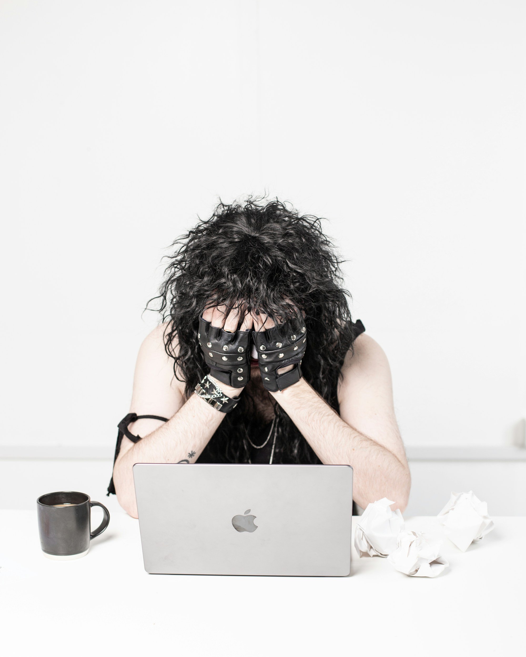 woman with fingerless gloves hides her face. In front of her is an Apple laptop, a cup of coffee, and a tangle of wires.