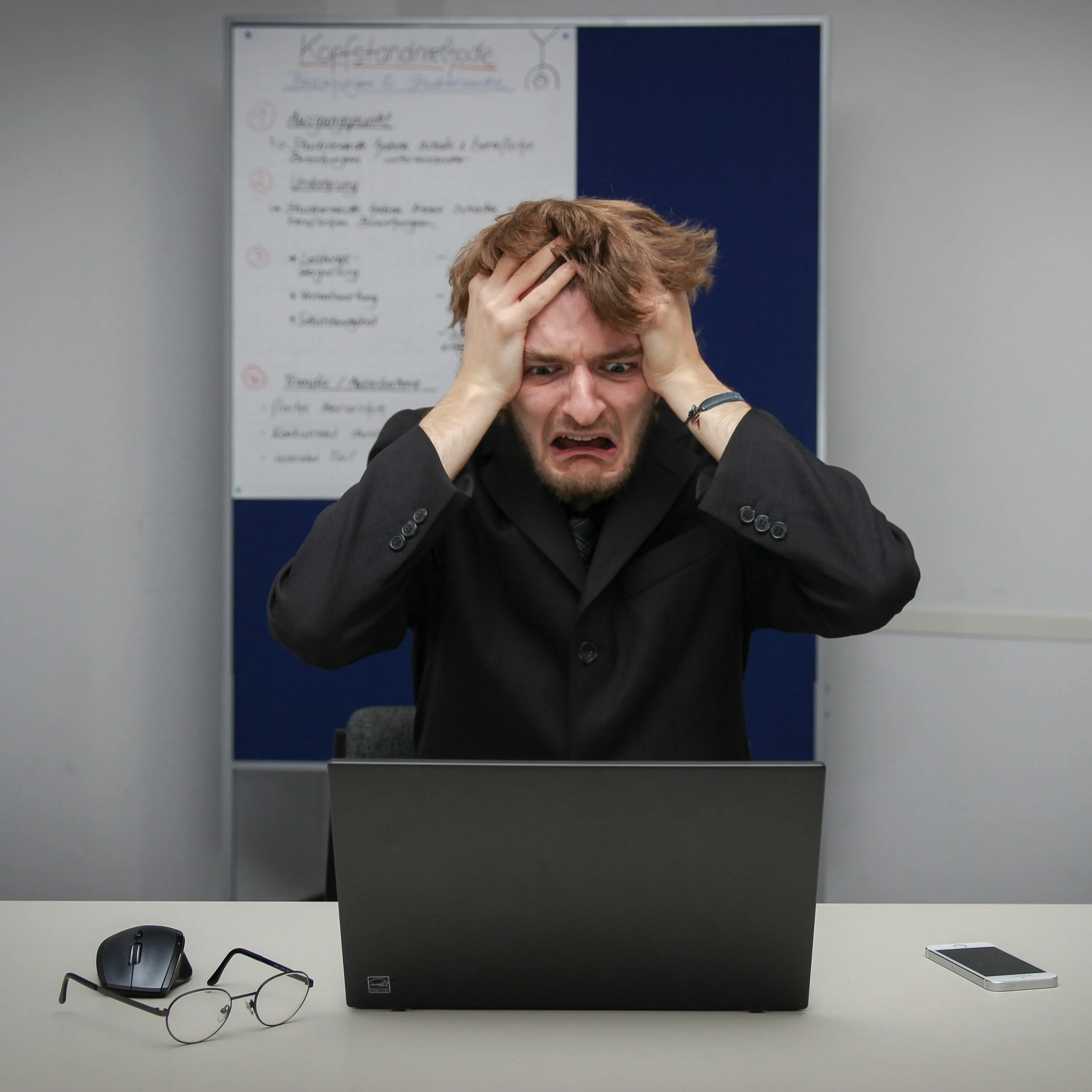 Male office worker with a frustrated expression holds his head in his hands. In front of him is a laptop, a phone, and his glasses. Behind hin is a board with indistinct writing on it.