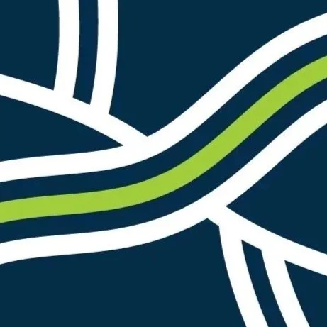 A woman with short, wavy, light brown hair smiling in front of painted landscapes, one with snow-covered trees and another with a blue sky and water.