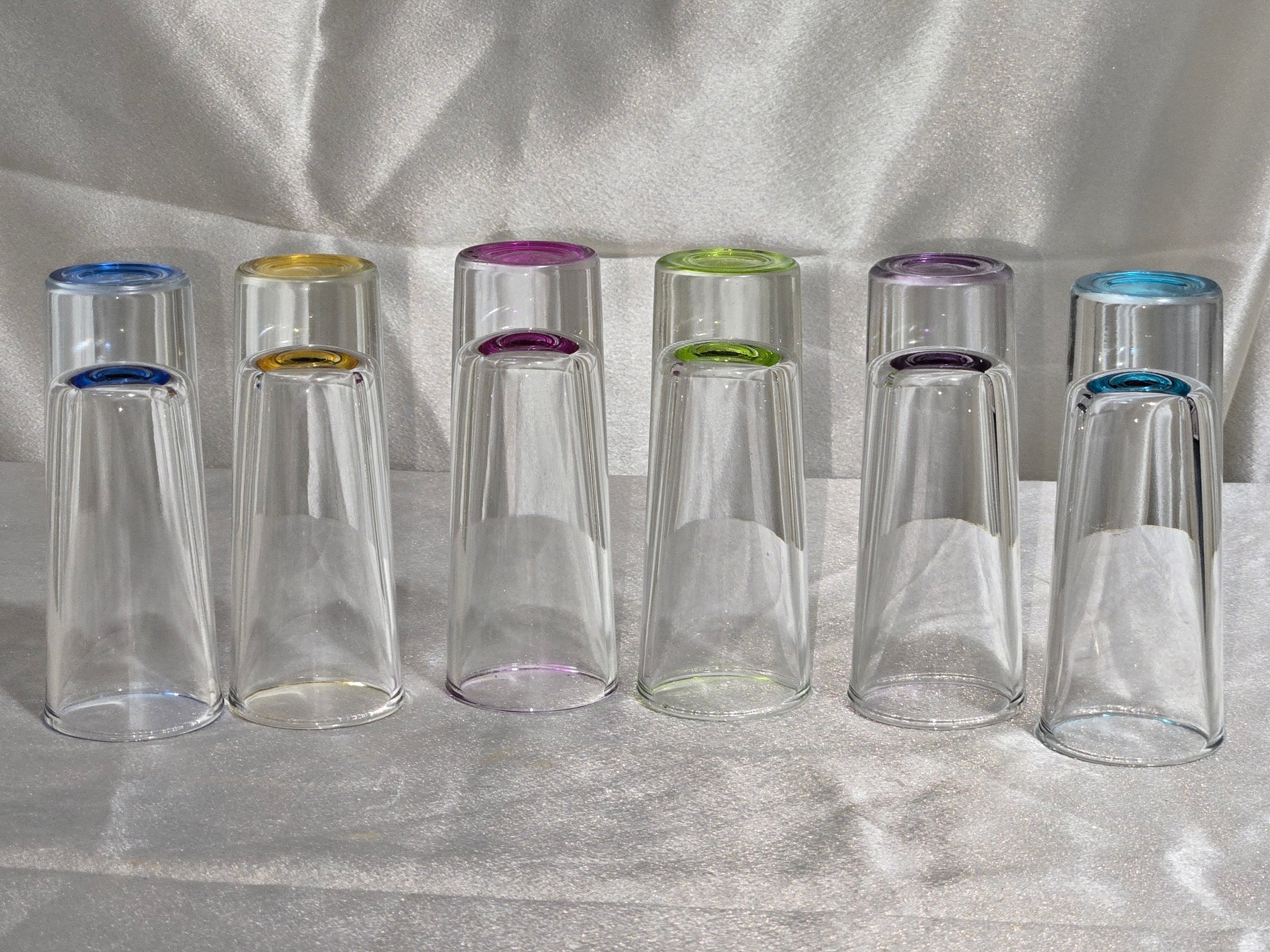 Set of six tall clear vintage shot glasses with colorful glass bases arranged in a row on white fabric