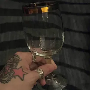 A hand holding a clear wine glass filled with water, tattoos and rings visible on fingers, against a dark striped background.