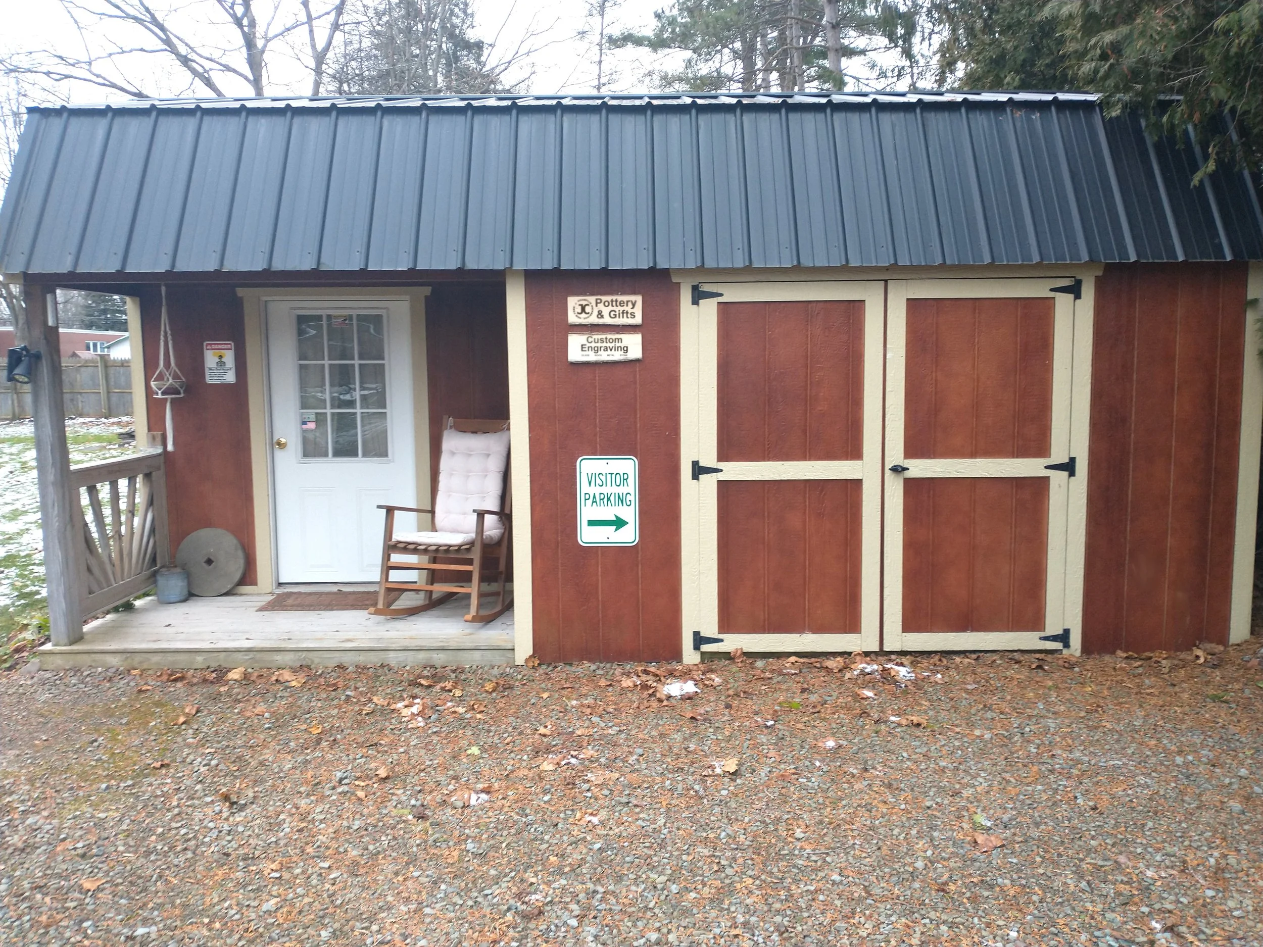 Red shed with cream trim, black metal roof, white door, and windows. A sign indicating visitor parking with right arrow, and two plaques reading 'Pottery & Gifts' and 'Custom Engraving'. A cushioned rocking chair on a small porch, and next to it a round stone and small bucket. Trees and a fence in the background.