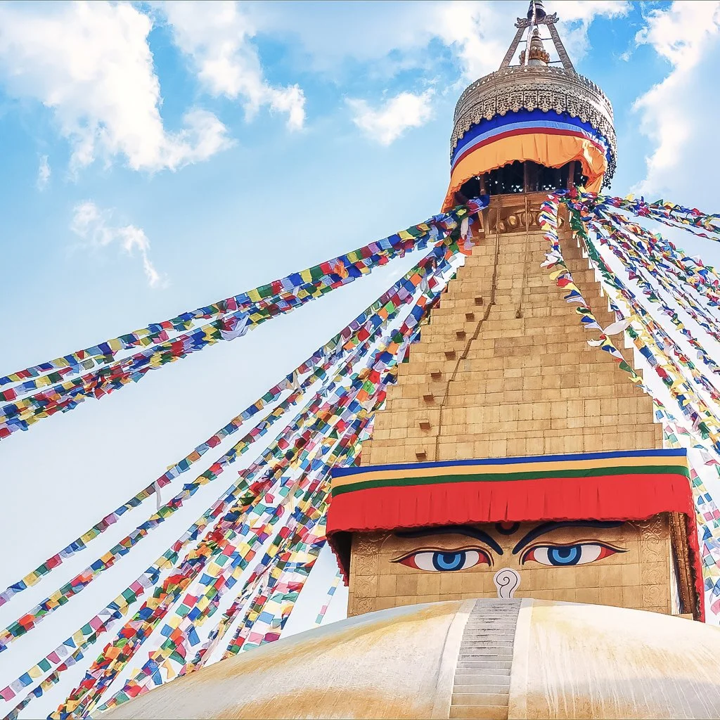 A tall Buddhist stupa with eye symbols at the base, decorated with colorful prayer flags against a blue sky with some clouds.