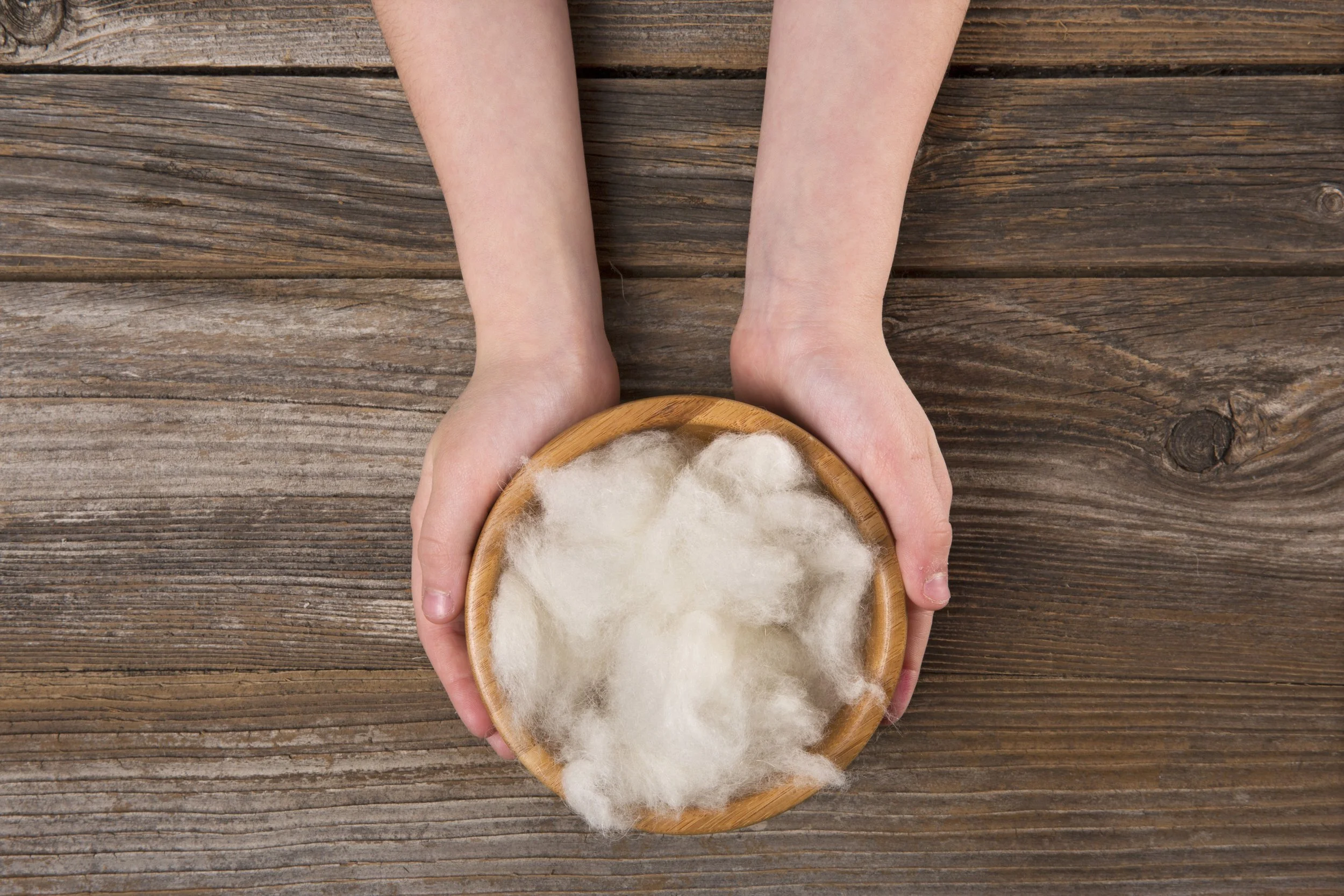 Hands holding a small wooden bowl filled with white fiber material, placed on a weathered wooden surface.