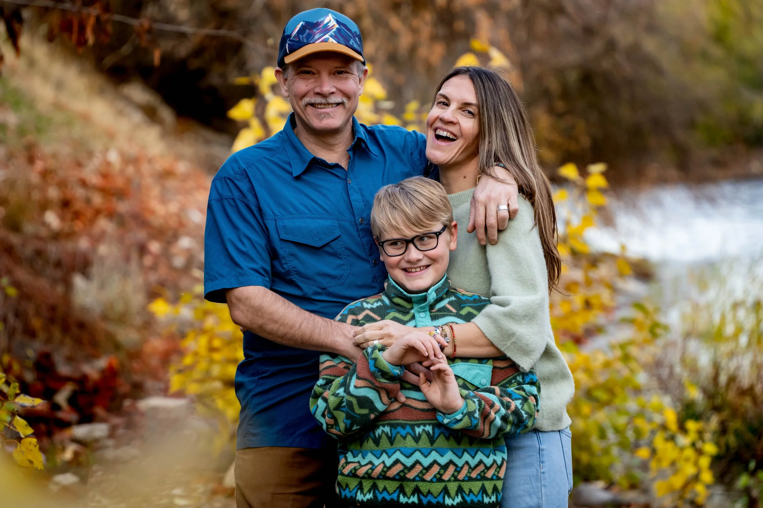 A happy family of three, a man, woman, and young boy, outdoors during autumn, embracing and smiling at the camera with fall foliage and a stream in the background.