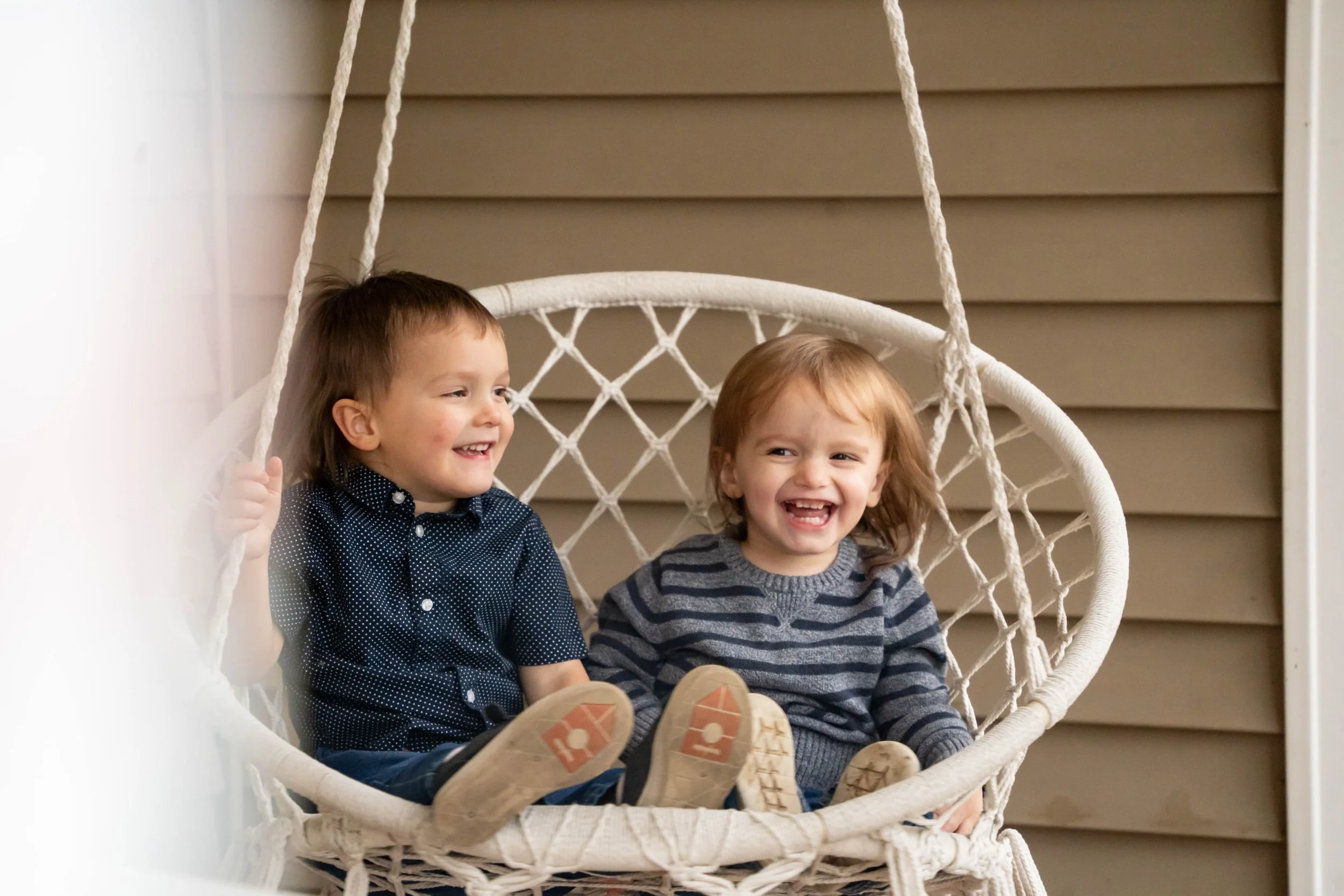 Two young children, a boy and a girl, sitting on a hanging chair outdoors, smiling and laughing.