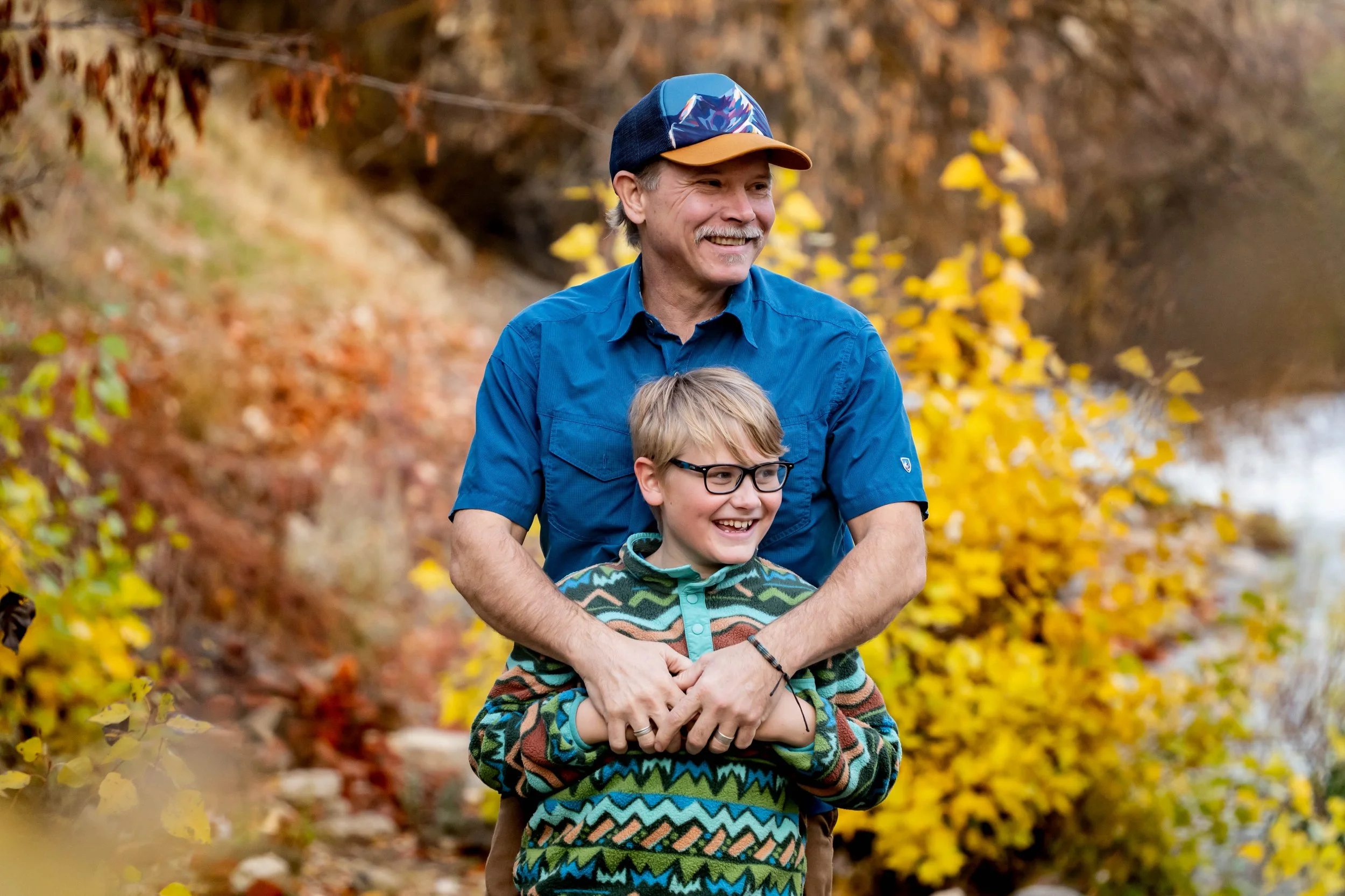 A smiling man and boy outdoors in autumn, with the man hugging the boy from behind in a wooded area with yellow and orange leaves.