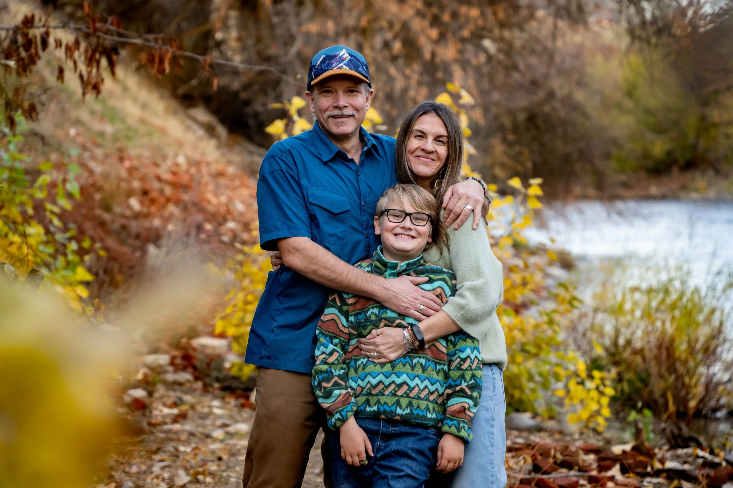 A family of three smiling and hugging outdoors in fall next to a river, with autumn leaves and trees in the background.