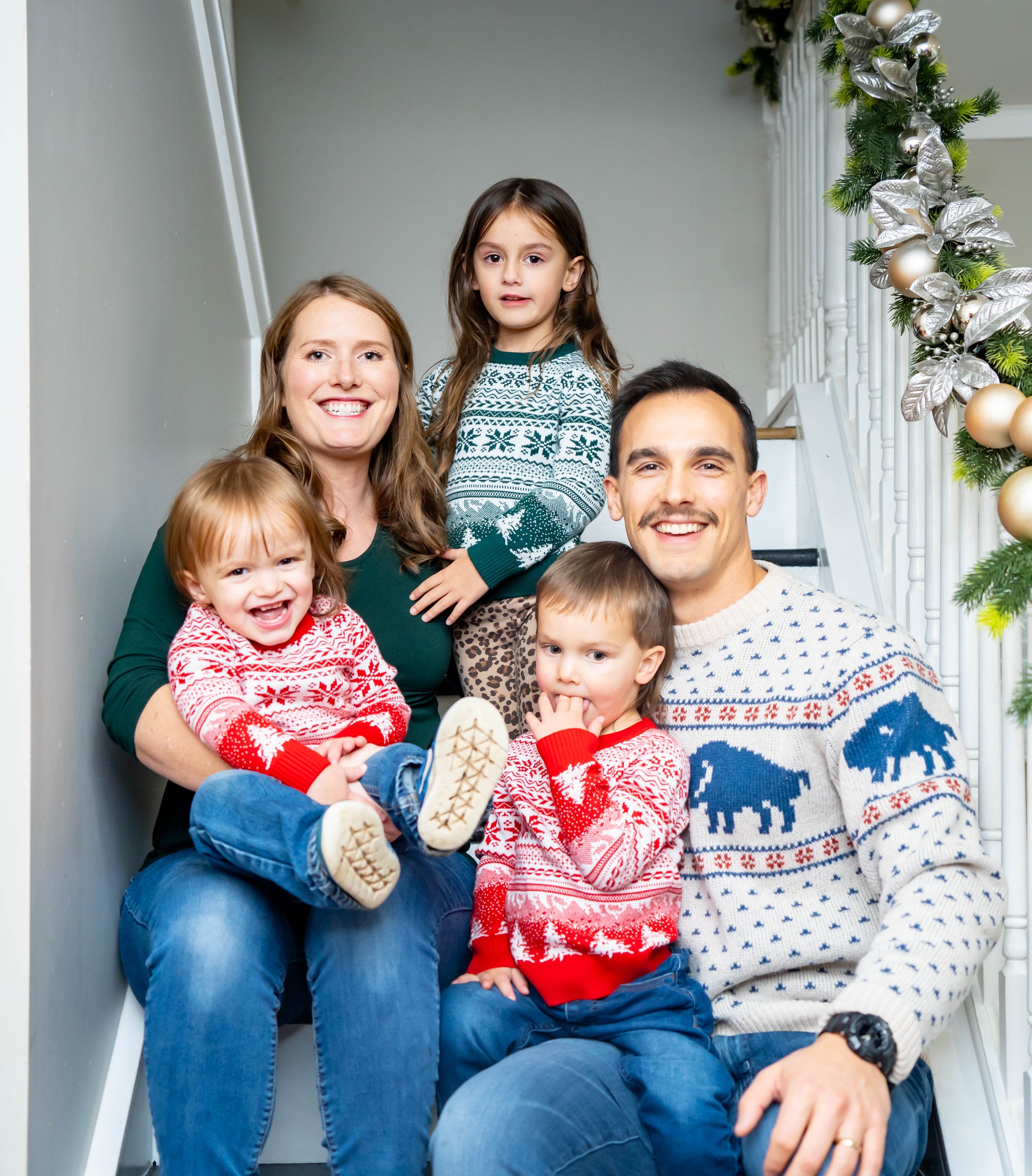 A family of six posing on a staircase decorated for Christmas, wearing holiday sweaters, with a Christmas garland on the side.
