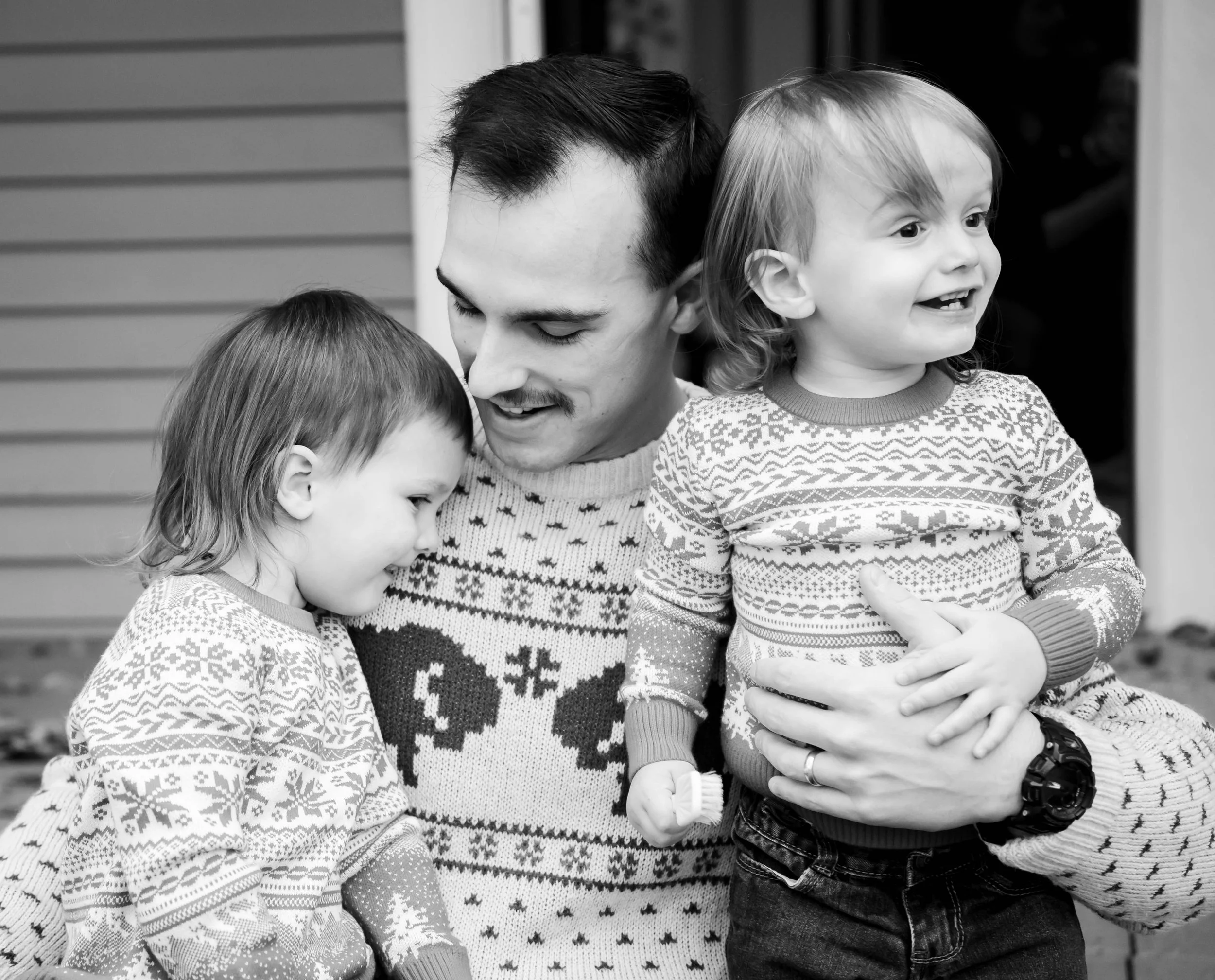 A man with two young girls, all wearing Christmas sweaters, outdoors near a house. The man is smiling and holding one girl, who is also smiling, while the other girl is leaning close with her eyes closed.
