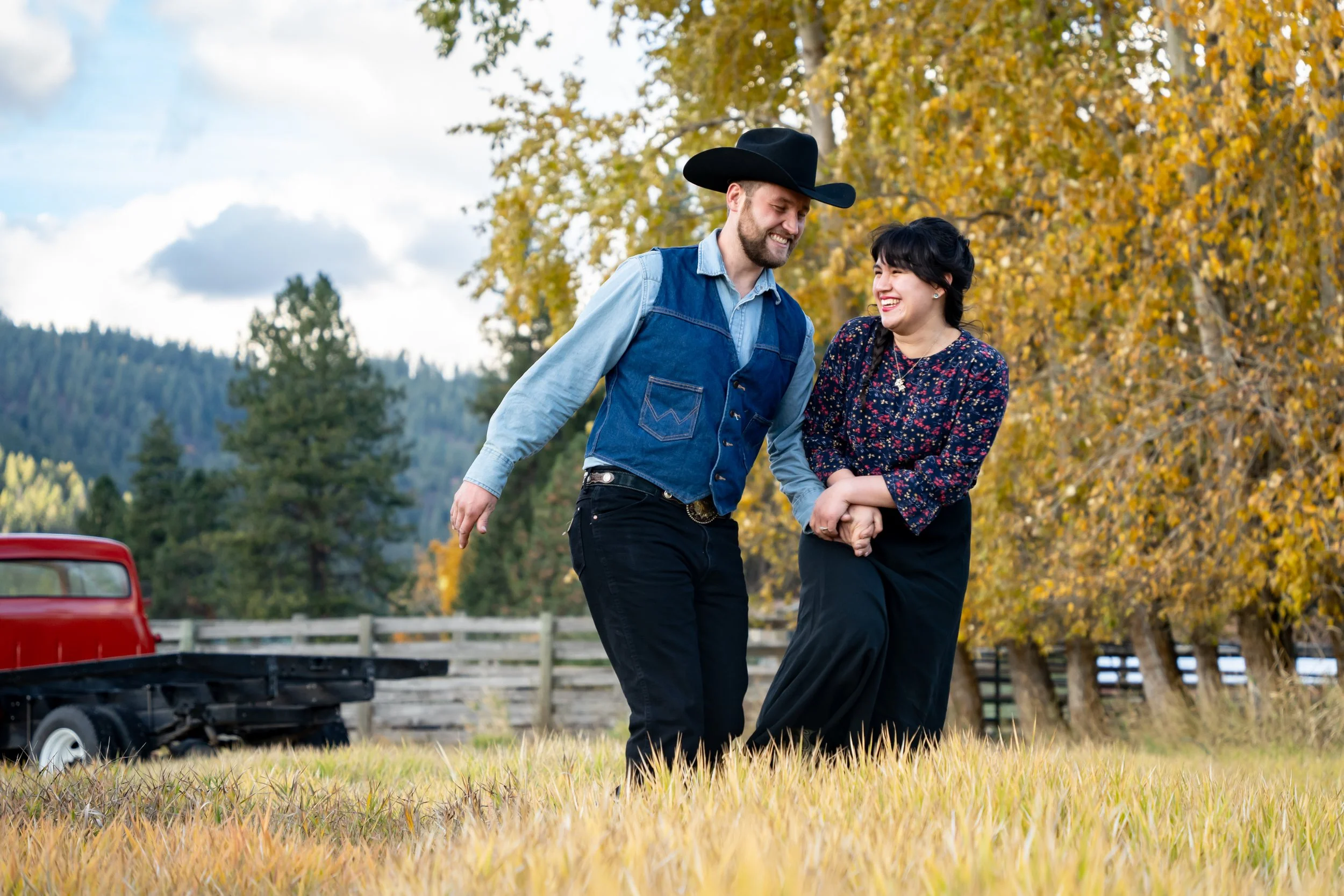 A happy couple walking hand in hand through a grassy field with autumn trees in the background, smiling at each other.