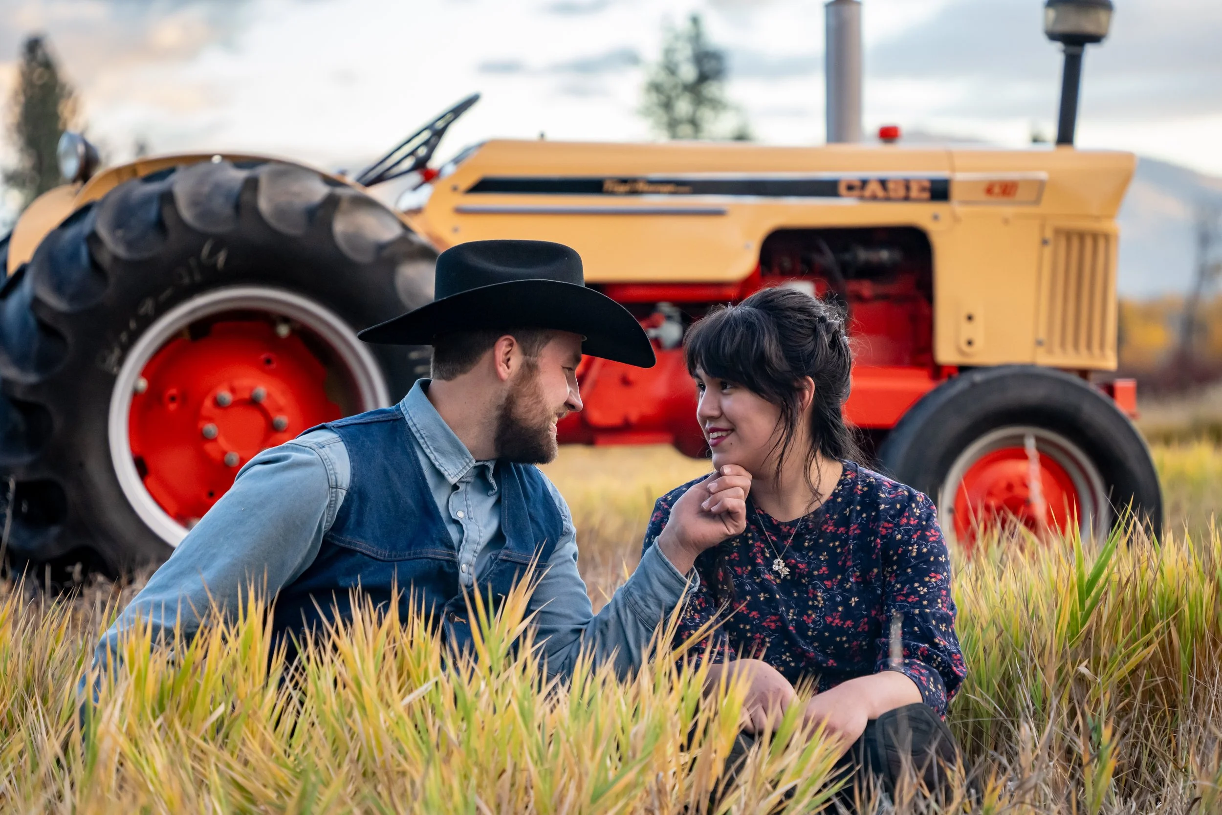 A man and woman sitting in a field of tall grass with a yellow tractor behind them, appearing to share an intimate moment.